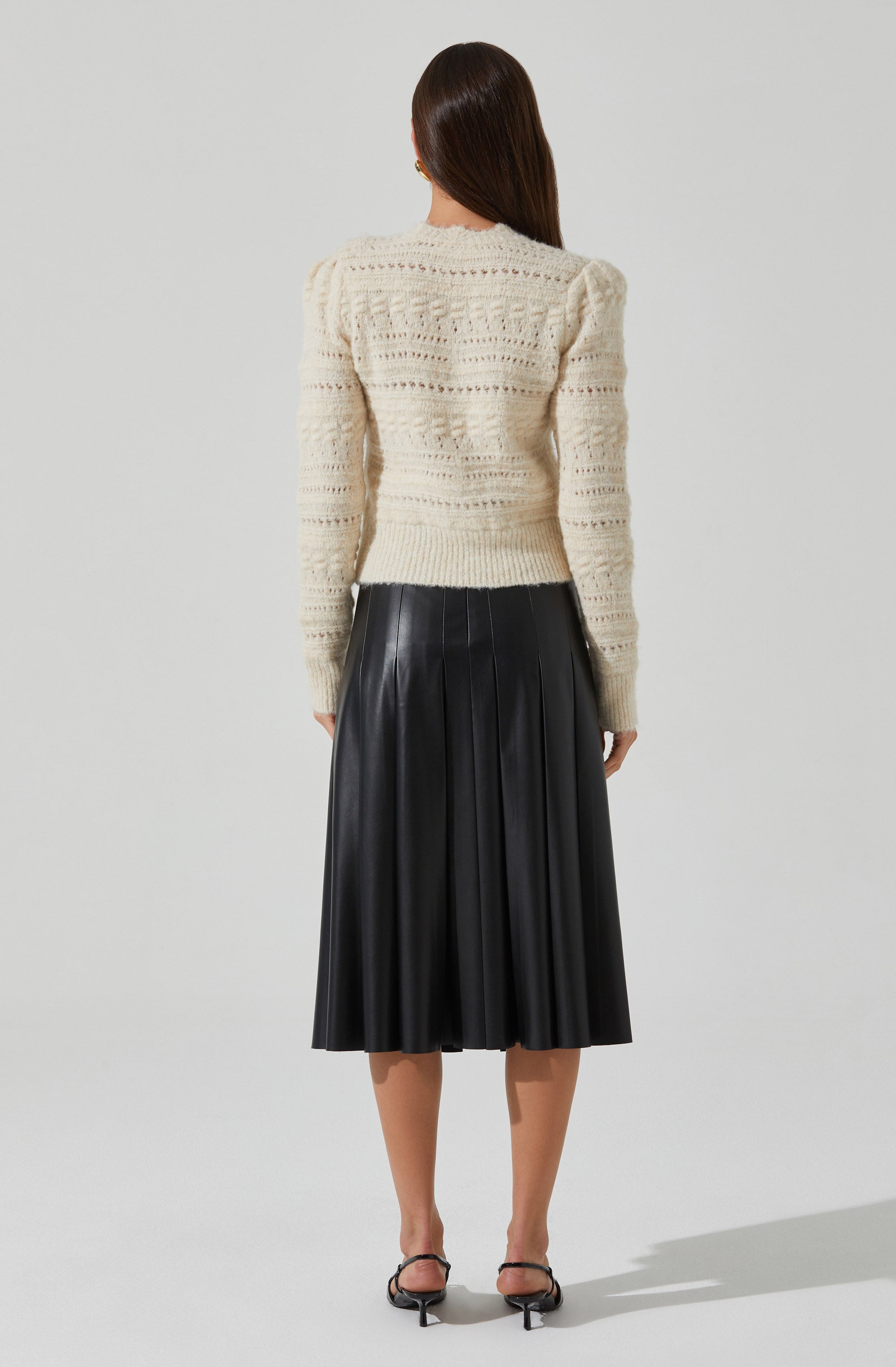 A woman is wearing a cream-colored, long-sleeved, knitted sweater with a textured pattern. She pairs it with a high-waisted, black pleated midi skirt that has a shiny leather appearance. She is also wearing black open-toe, ankle-strap high-heeled shoes.