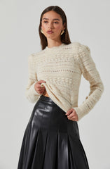 A woman is wearing a cream-colored, knit sweater with a textured, open weave pattern. She is also wearing a high-waisted, black leather skirt with wide pleats. She has large, gold statement earrings and a gold ring on her finger.