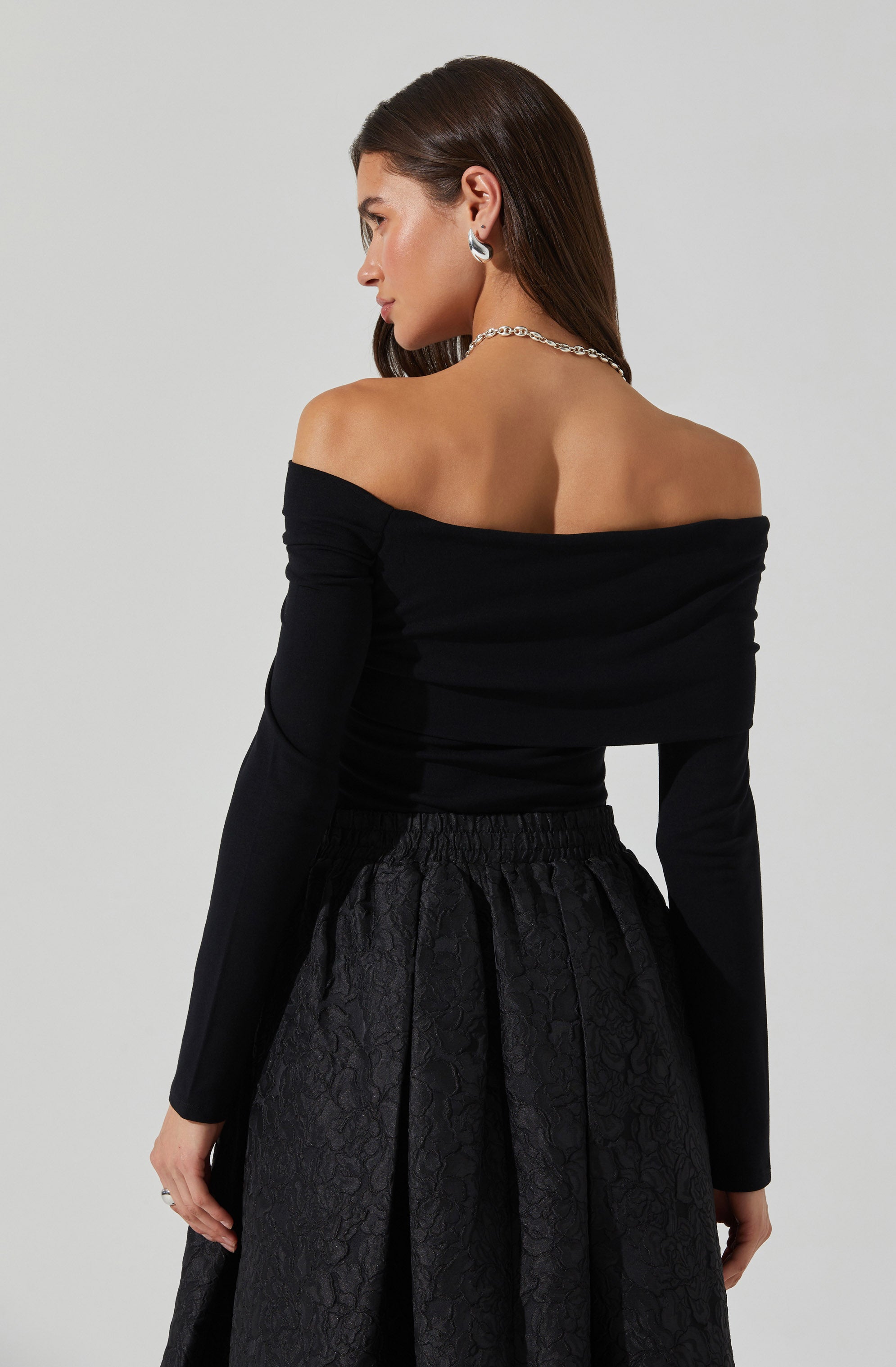 A woman is wearing an off-the-shoulder black top with long sleeves. She pairs it with a high-waisted, textured black skirt that features a subtle floral pattern. She accessorizes with silver hoop earrings and a chain necklace.