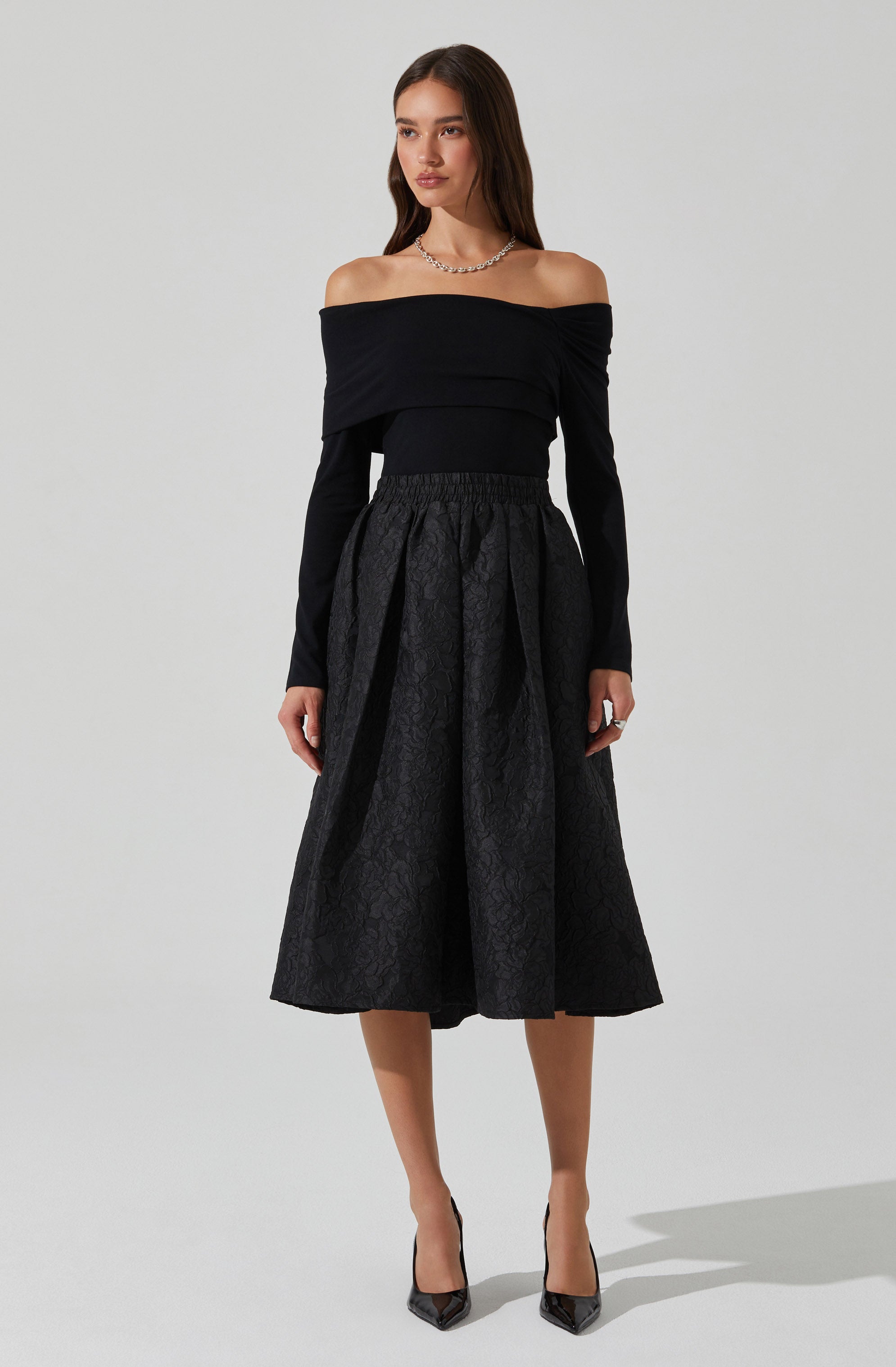 A woman is wearing a black off-the-shoulder top with long sleeves, paired with a high-waisted, midi-length skirt that has a subtle textured pattern. She completes the outfit with pointed-toe heels.