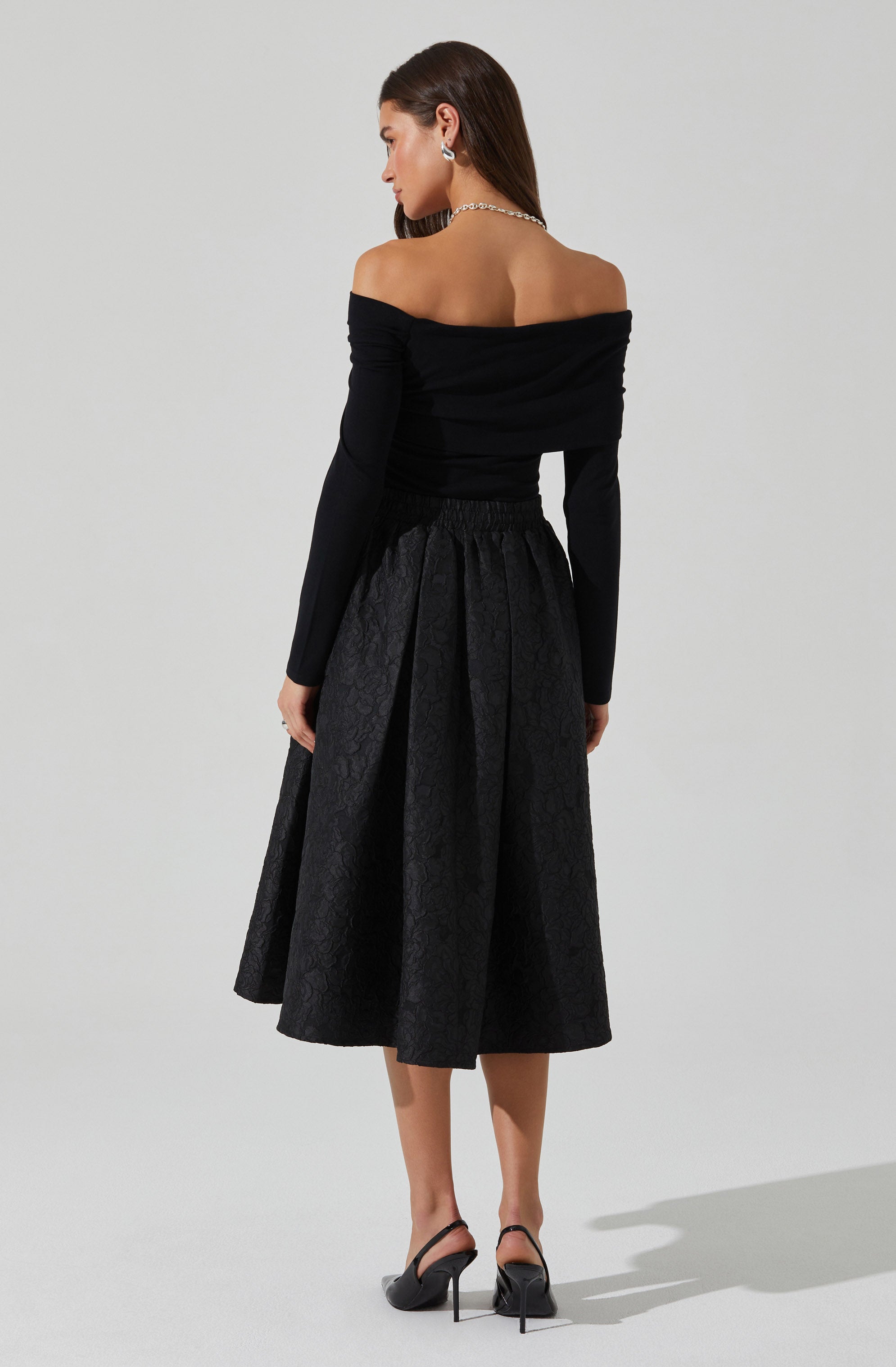 A woman is wearing an off-the-shoulder black top with long sleeves. She pairs it with a high-waisted, midi-length black skirt that has a subtle textured pattern. She completes her outfit with black high-heeled shoes that have ankle straps.