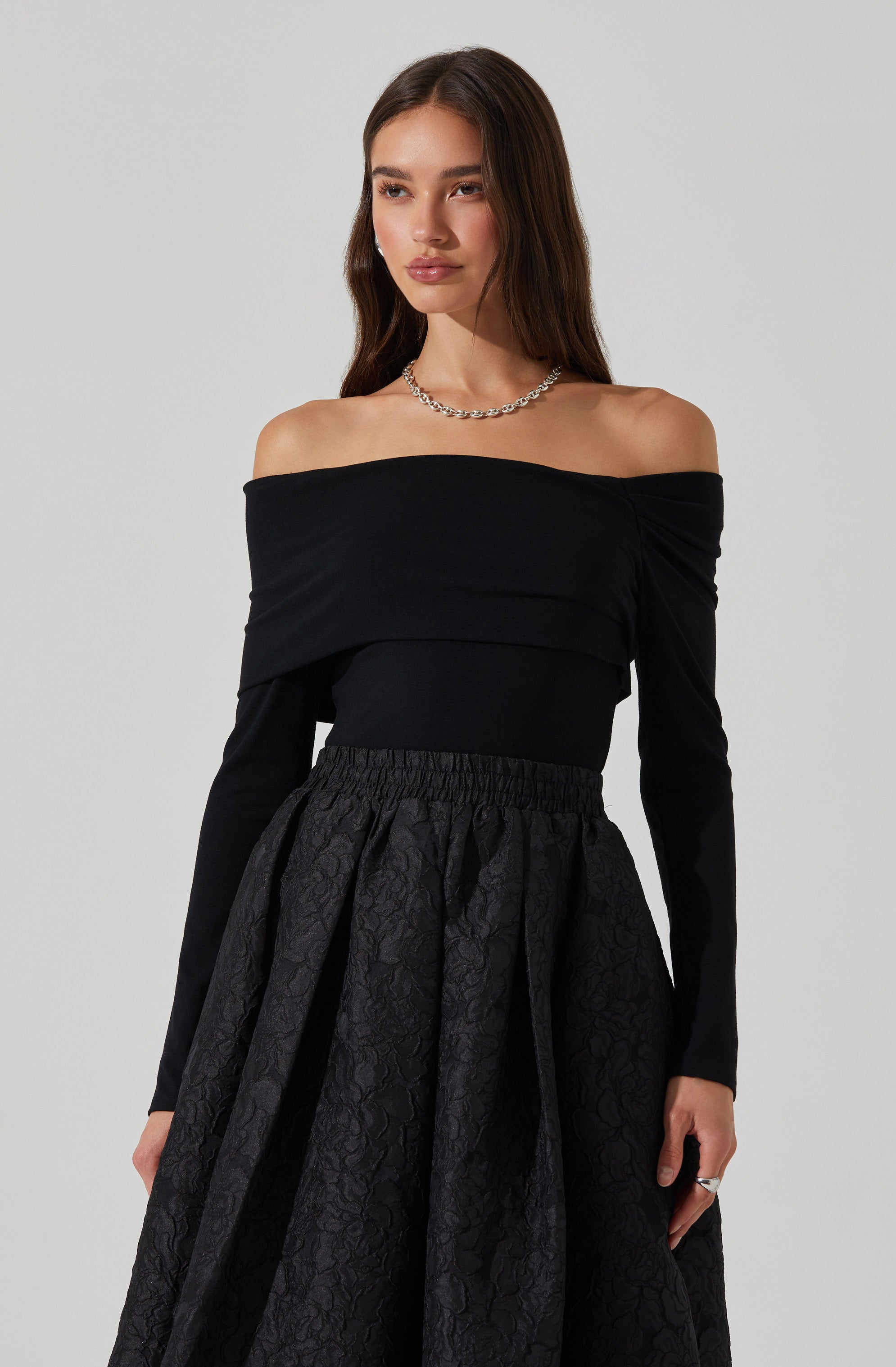 A woman is wearing an off-the-shoulder black top with long sleeves and a fitted waistband. She pairs it with a high-waisted, full skirt that has a textured, floral pattern and a gathered, voluminous silhouette. She accessorizes with a silver chain necklace and a ring on her right hand.