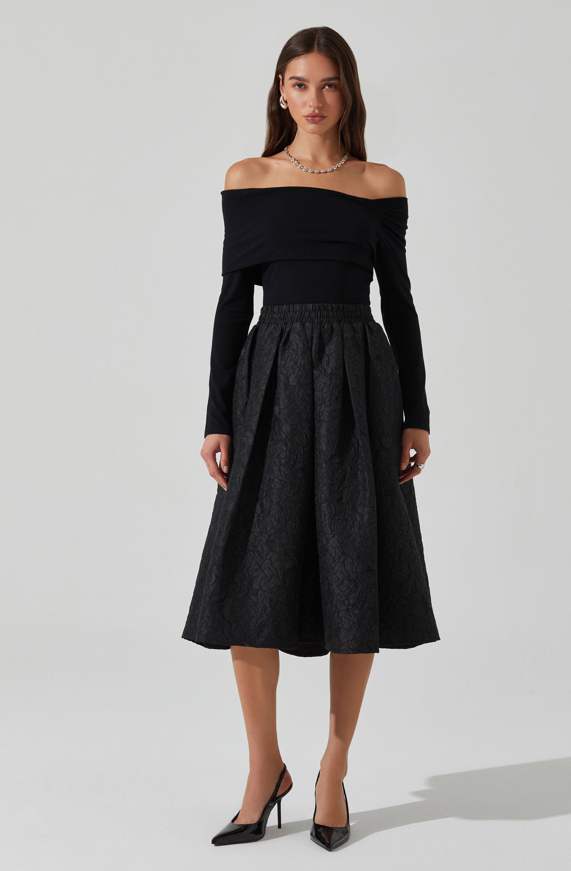 The woman is wearing an off-the-shoulder black top with long sleeves, paired with a high-waisted, full-length black skirt that features a textured, patterned fabric. She accessorizes with a necklace, earrings, and black high-heeled shoes.