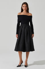 The woman is wearing an off-the-shoulder black top with long sleeves, paired with a high-waisted, full-length black skirt that features a textured, patterned fabric. She accessorizes with a necklace, earrings, and black high-heeled shoes.
