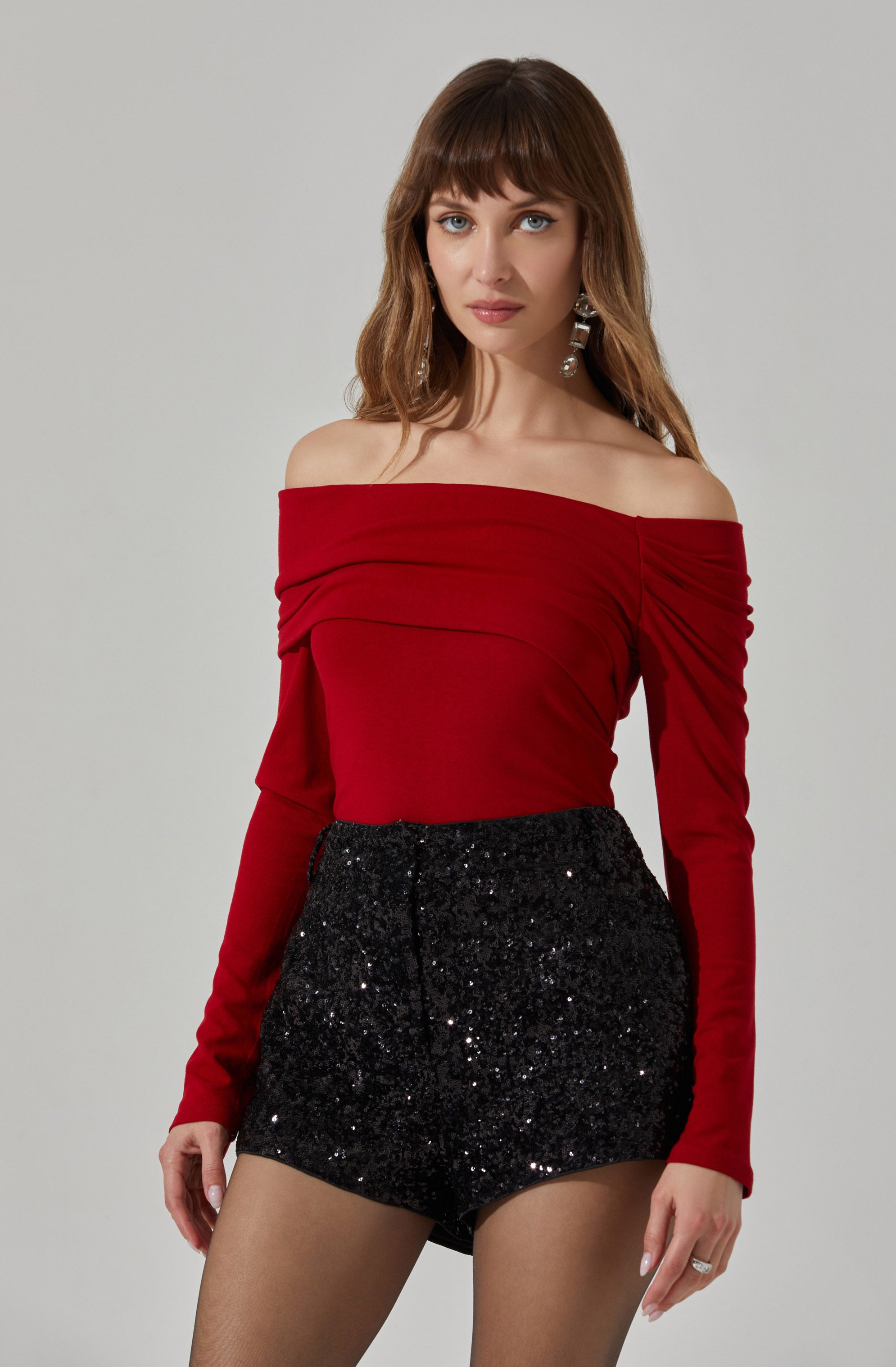 A woman is wearing a red off-the-shoulder top with long sleeves that have ruching details on the upper arms, and high-waisted black shorts covered in sequins that create a sparkly effect.
