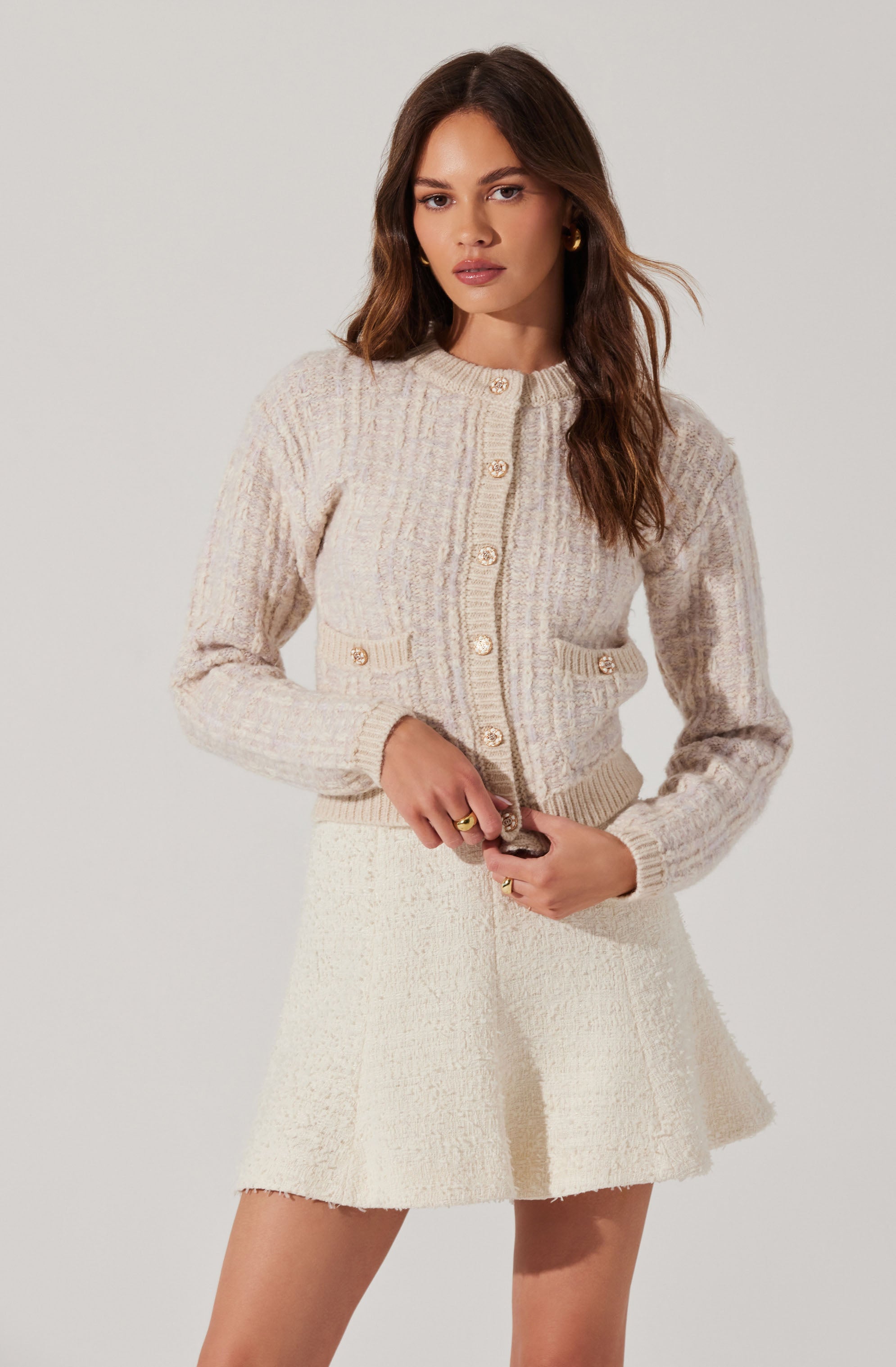 A woman is wearing a light-colored, textured knit cardigan with large buttons down the front, and two small decorative pockets on the front with matching buttons. She has paired it with a cream-colored, textured, A-line mini skirt.