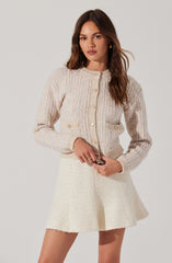 A woman is wearing a light-colored, textured knit cardigan with large buttons down the front, and two small decorative pockets on the front with matching buttons. She has paired it with a cream-colored, textured, A-line mini skirt.