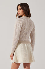 A woman is wearing a light beige, ribbed knit sweater with long sleeves and a round neckline. She pairs it with an off-white, textured, A-line skirt that has a slightly frayed hem, creating a soft and cozy look.