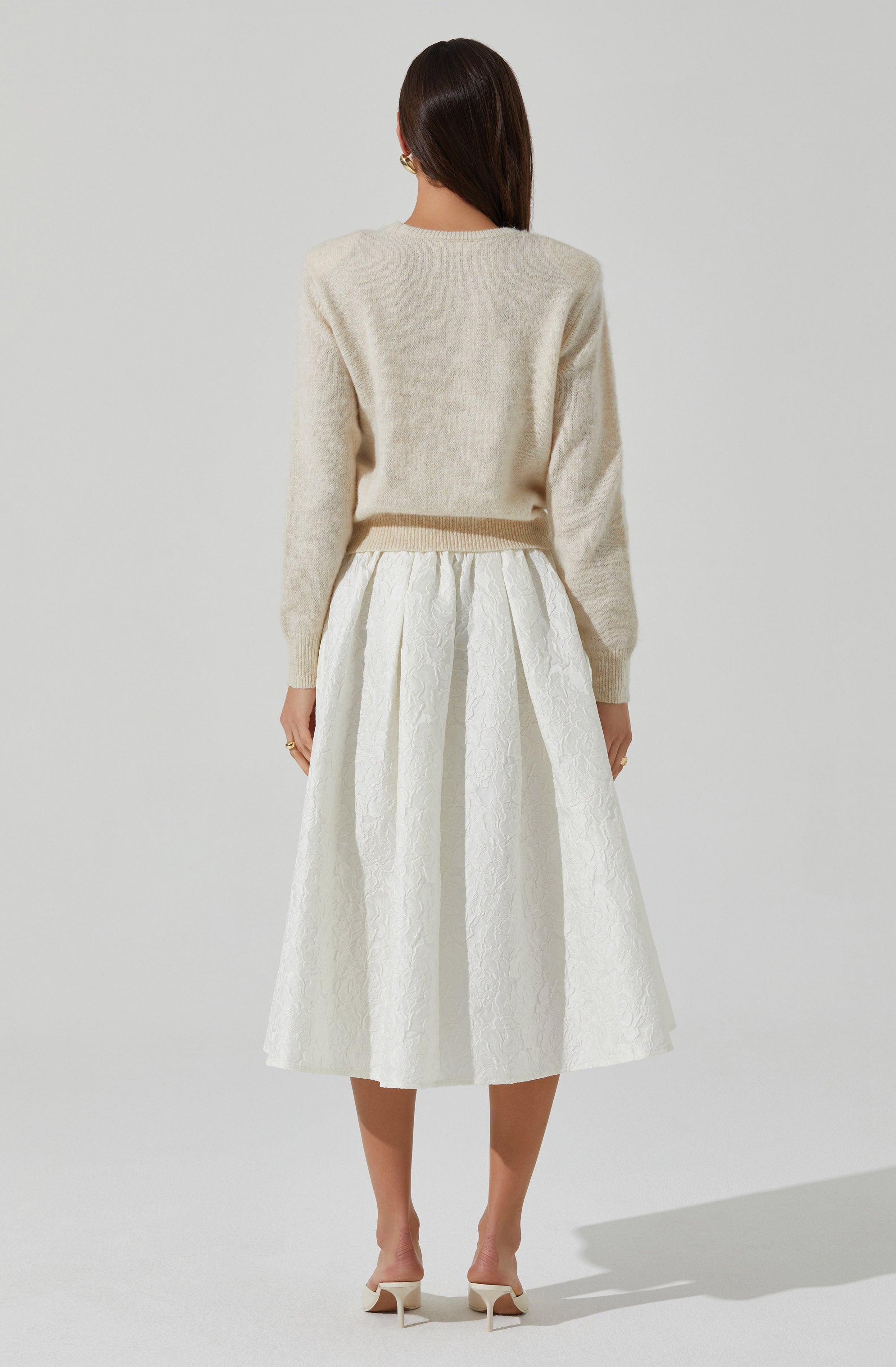 A woman is wearing a light cream, fuzzy knit sweater with long sleeves and a round neckline. She pairs it with a high-waisted, white midi skirt that features a textured, floral pattern and pleats. She completes her outfit with white, open-toe high-heeled shoes with ankle straps.