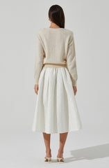 A woman is wearing a light cream, fuzzy knit sweater with long sleeves and a round neckline. She pairs it with a high-waisted, white midi skirt that features a textured, floral pattern and pleats. She completes her outfit with white, open-toe high-heeled shoes with ankle straps.