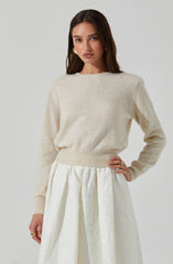 A woman is wearing a soft, cream-colored, knit sweater with a round neckline and long sleeves. She pairs it with a high-waisted, white textured skirt that features pleats and a subtle floral pattern.