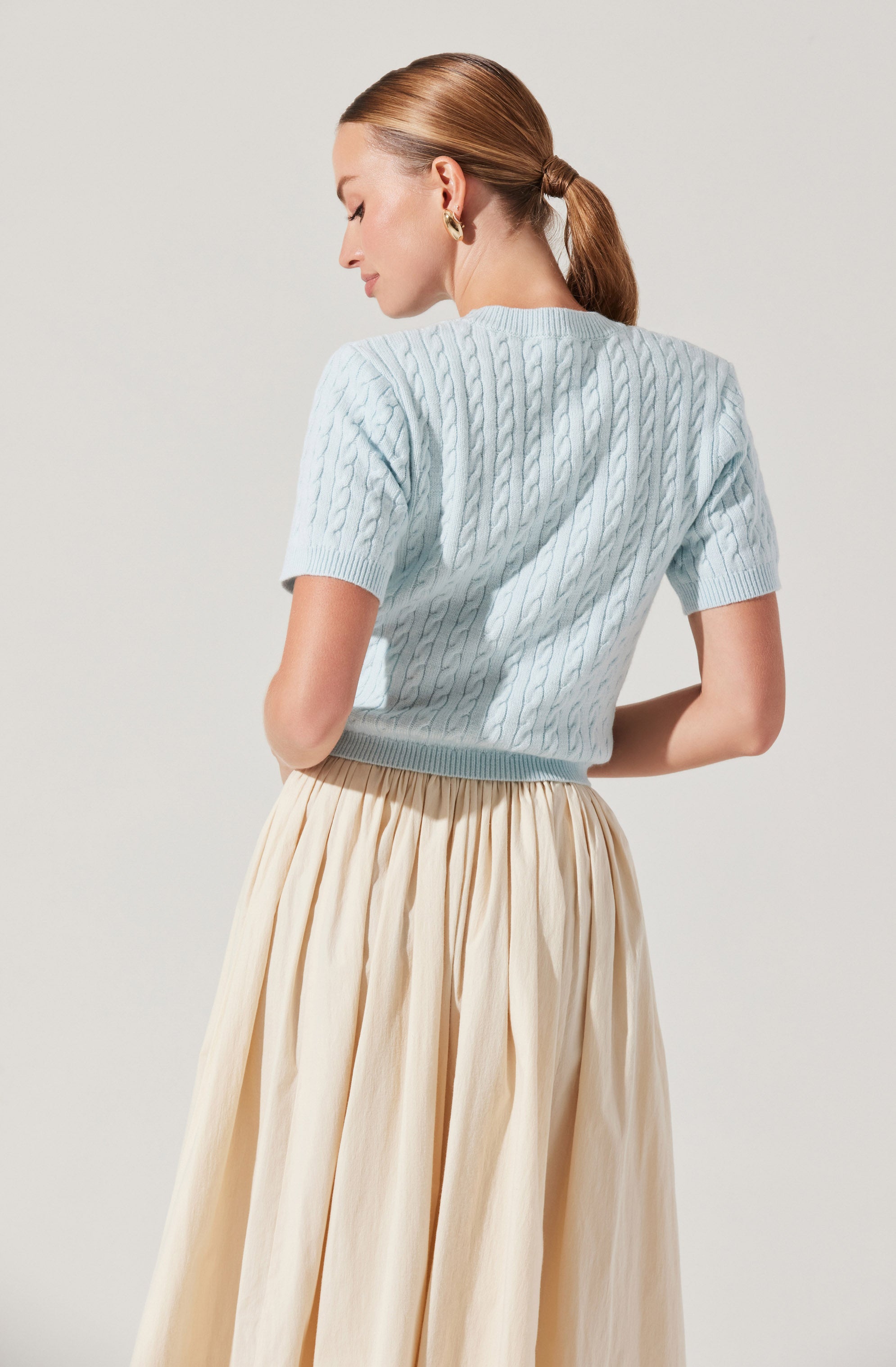 A woman is wearing a light blue, short-sleeved cable-knit sweater and a high-waisted, cream-colored, pleated skirt. The sweater has a textured, woven pattern, and she has her hair tied back in a low ponytail. She is turned slightly to the side with her eyes closed.