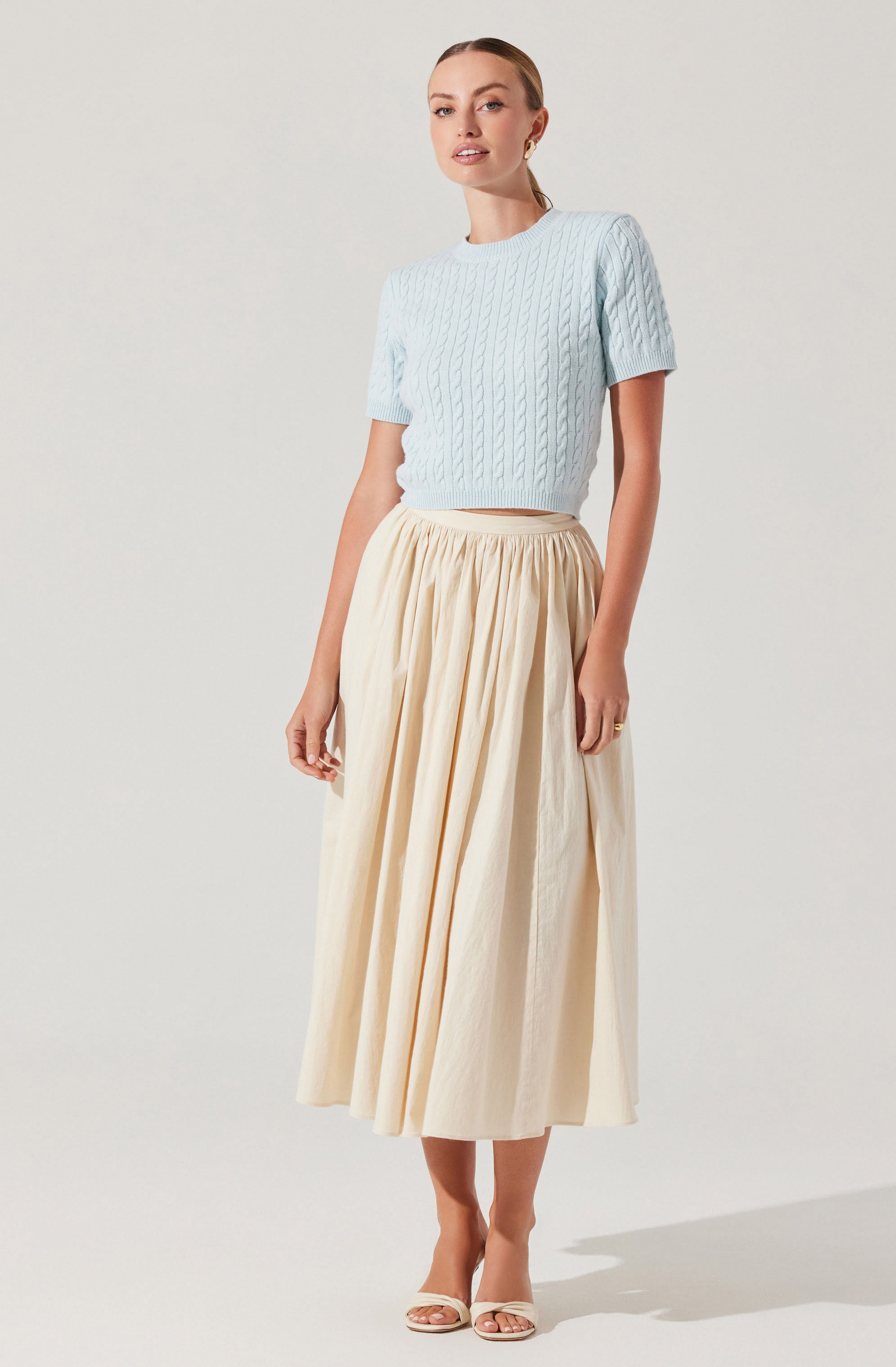 A woman is wearing a light blue, short-sleeved, cable-knit sweater with a round neckline. She pairs it with a high-waisted, full-length, cream-colored skirt that has a gathered, flowing design. She completes the outfit with open-toe, cream-colored heels.