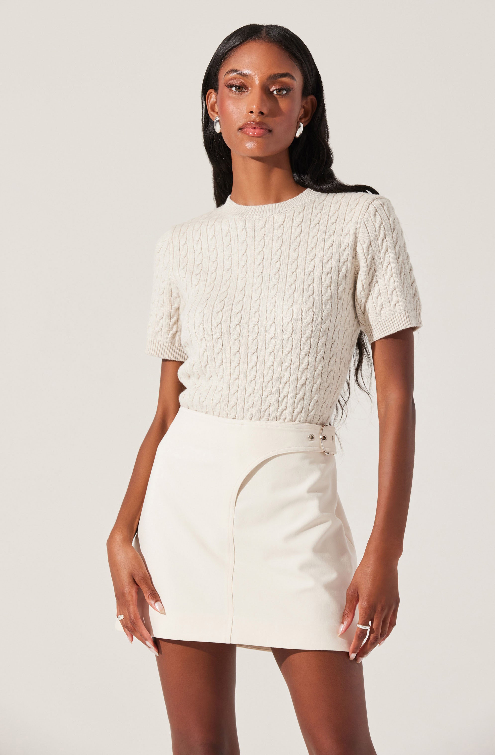 A woman is wearing a short-sleeved, cable-knit cream sweater and a high-waisted, cream skirt with a side slit and buckle detail.