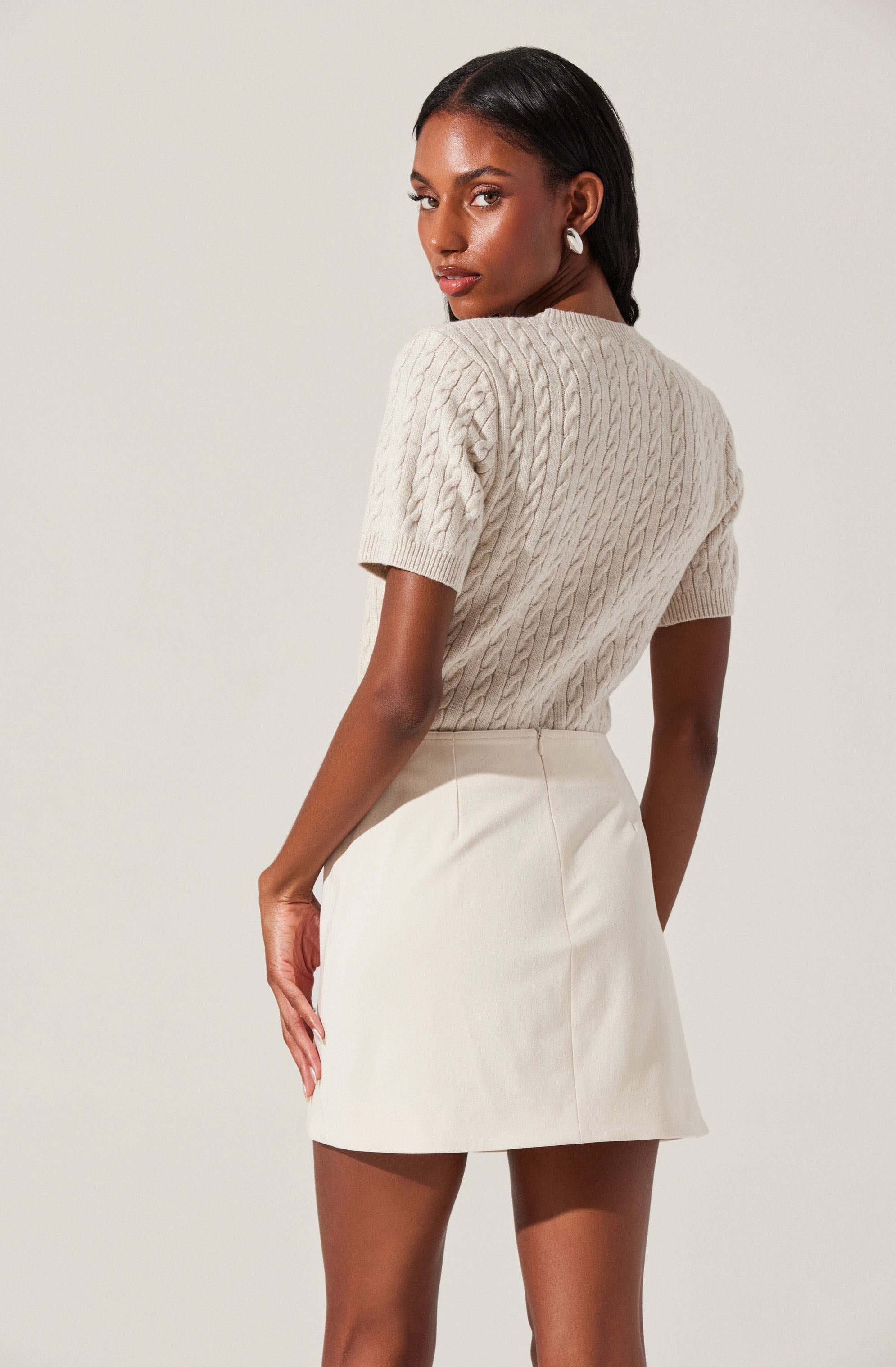 A woman is wearing a beige, cable-knit, short-sleeve sweater with a round neckline. She pairs it with a high-waisted, cream-colored, A-line mini skirt.