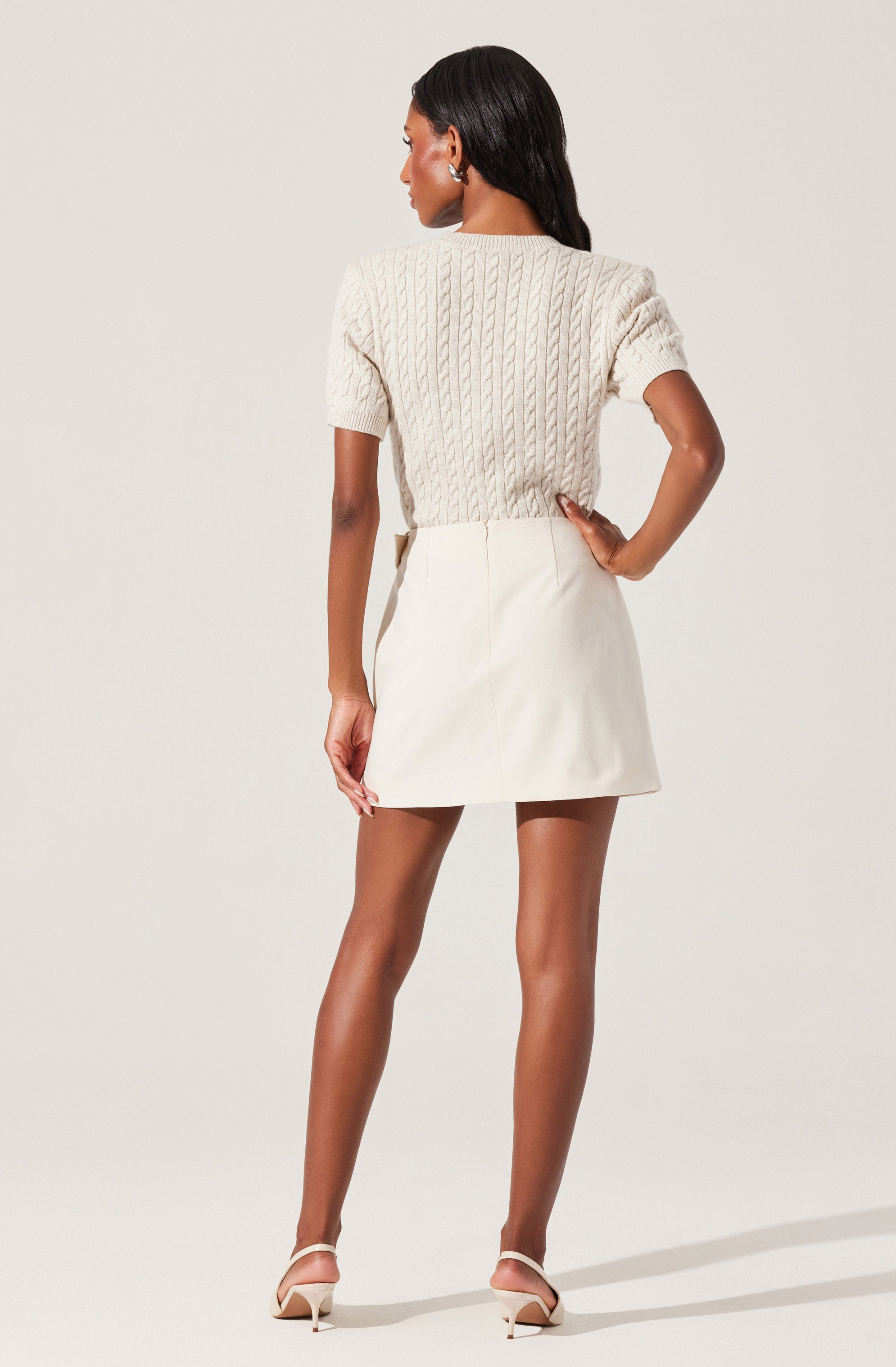 A woman is wearing a cream-colored, short-sleeved, cable-knit sweater paired with a matching cream-colored A-line mini skirt. She is also wearing nude high-heeled sandals.