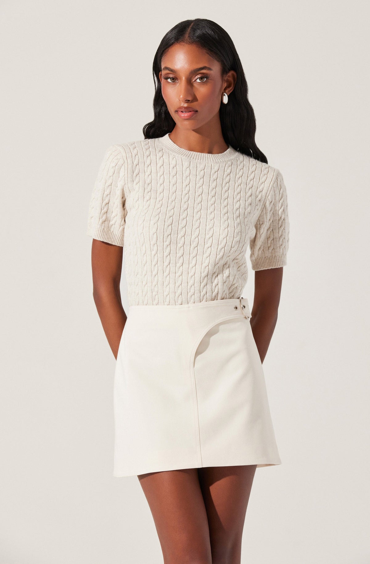 A woman wearing a cream-colored, short-sleeved cable knit sweater with a round neckline. She has on a high-waisted, cream-colored skirt with an asymmetrical zipper detail on the side. She accessorizes with large pearl earrings.