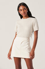 A woman is wearing a short-sleeved, cream-colored cable knit sweater with a round neckline. She pairs it with a high-waisted, cream-colored mini skirt featuring a small slit and button details on the waistband.