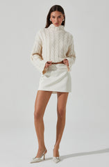 A woman is wearing an oversized, cream-colored cable-knit sweater with a high neckline and long, loose sleeves. She pairs it with a short, white, high-waisted skirt and white high-heeled shoes. She accessorizes with large, white hoop earrings.