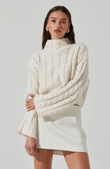 A woman is wearing a cream-colored, chunky knit sweater with a high neckline and cable-knit pattern. She also has on a white, high-waisted, A-line mini skirt. She accessorized with hoop earrings and a ring on her left hand.