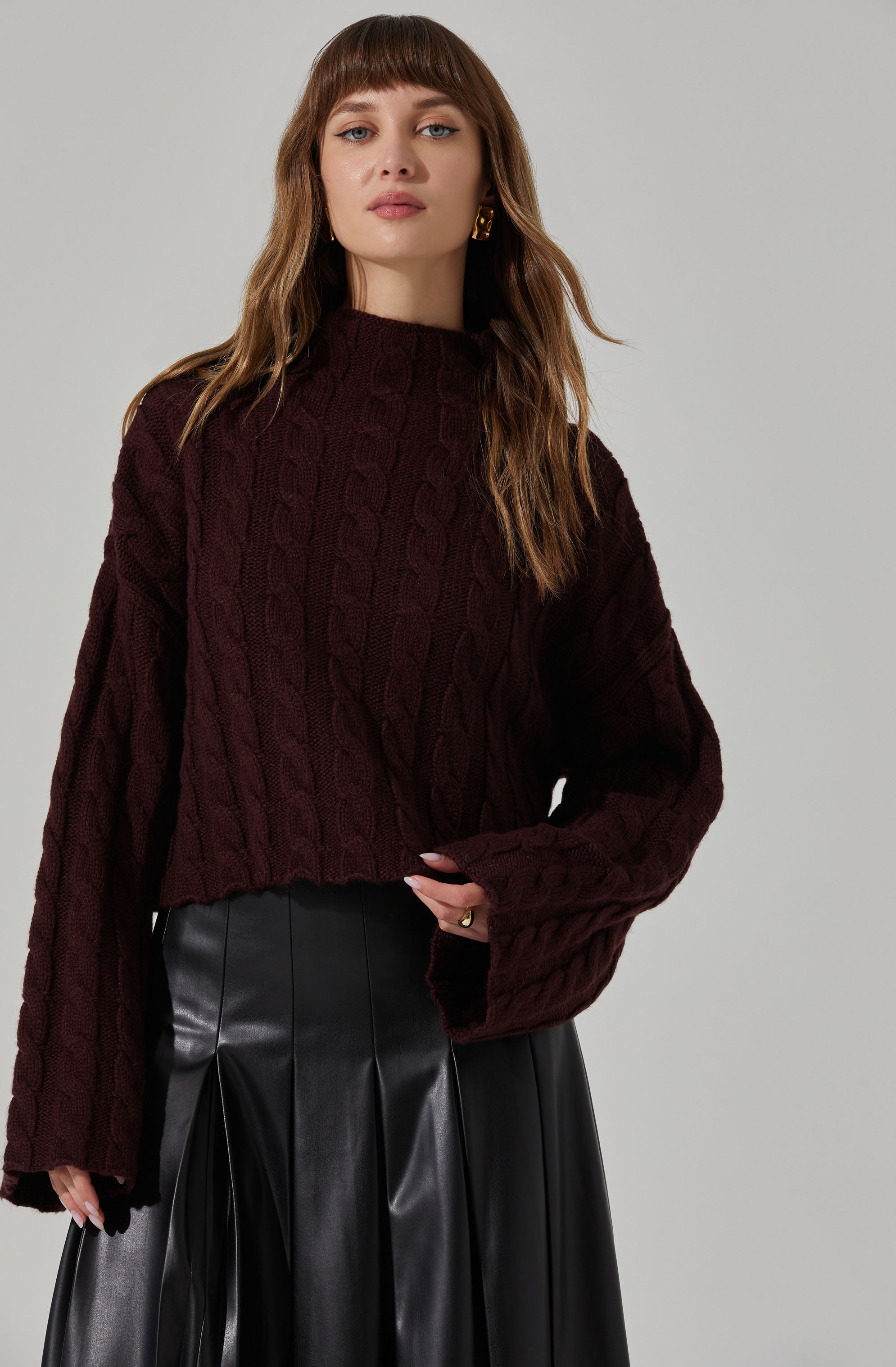A woman is wearing a dark burgundy, chunky knit sweater with a cable pattern. The sweater has a loose fit with long, wide sleeves. She pairs it with a black, high-waisted, shiny leather or faux leather skirt that has pleats. She also has gold hoop earrings and a ring visible on her left hand.