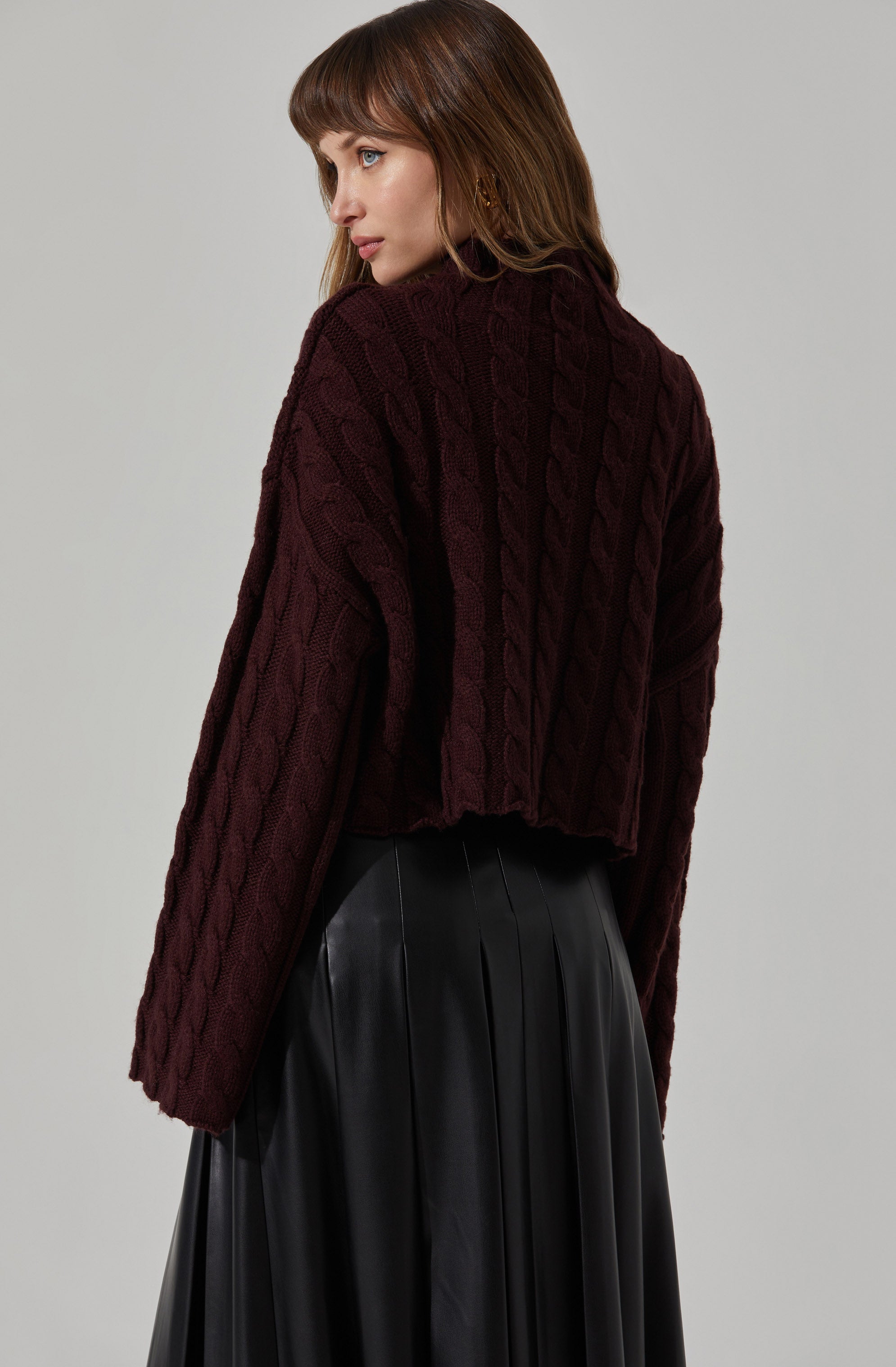 A woman is wearing a dark maroon, cable-knit, long-sleeved sweater with a high neckline. She is also dressed in a black, shiny, pleated skirt.