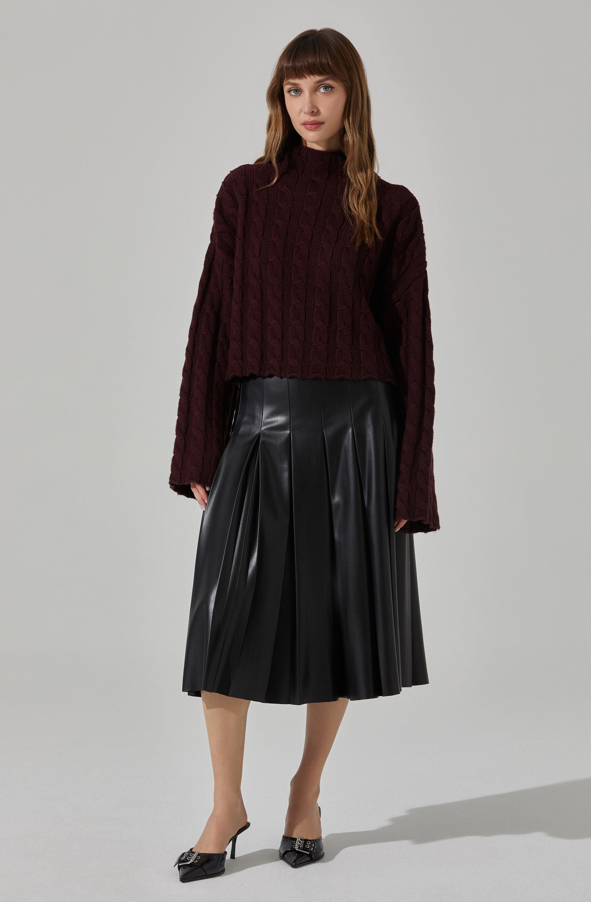 The woman is wearing a dark burgundy, chunky cable-knit sweater with a high neckline and long sleeves. She pairs it with a black, pleated, leather midi skirt that falls below the knees. She is also wearing black, pointed-toe high heels with small bow embellishments on the front.