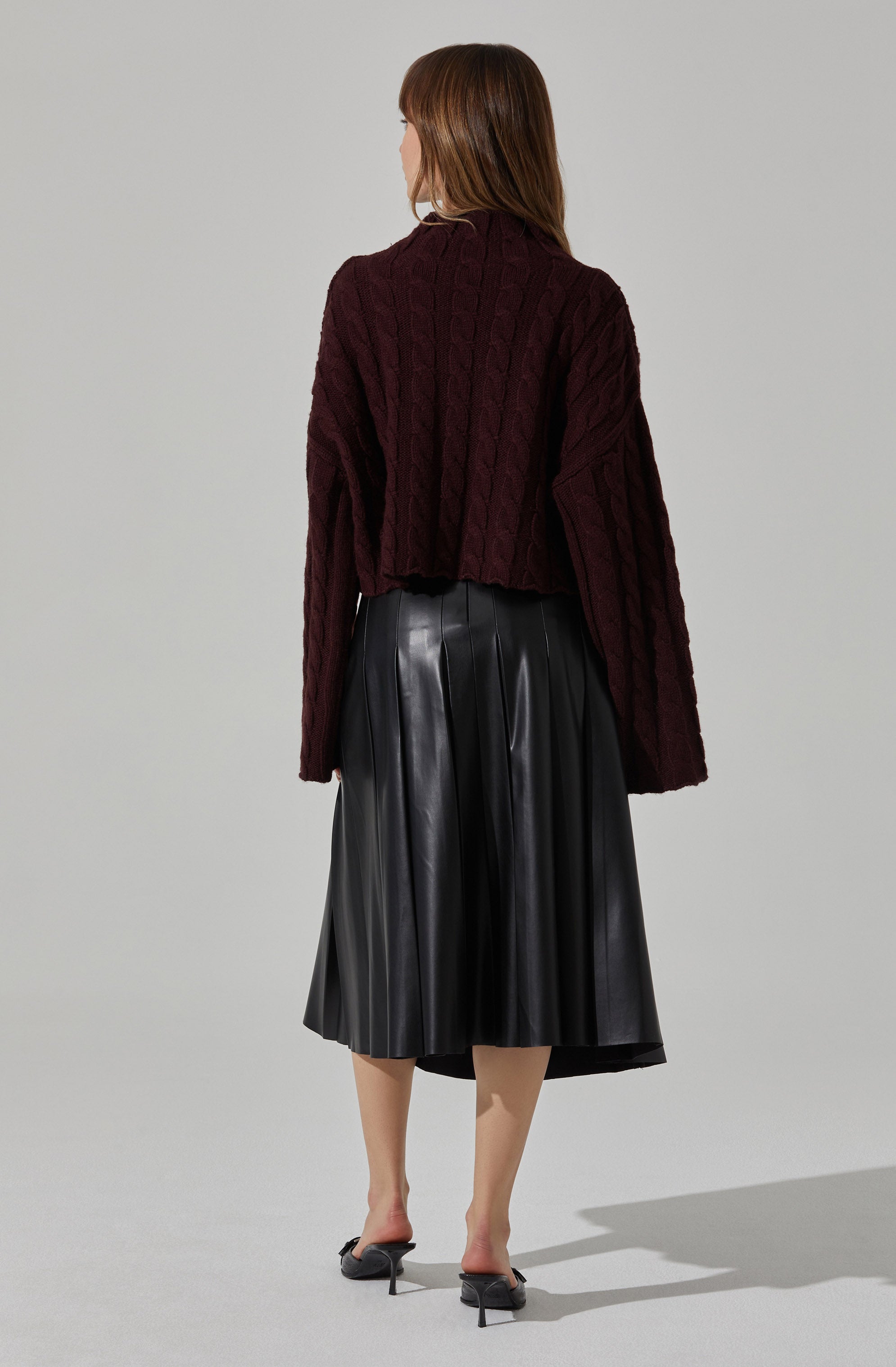 A woman is wearing a dark red, chunky knit sweater with a cable pattern, paired with a black, shiny, pleated midi skirt. She is also wearing black high-heeled sandals.