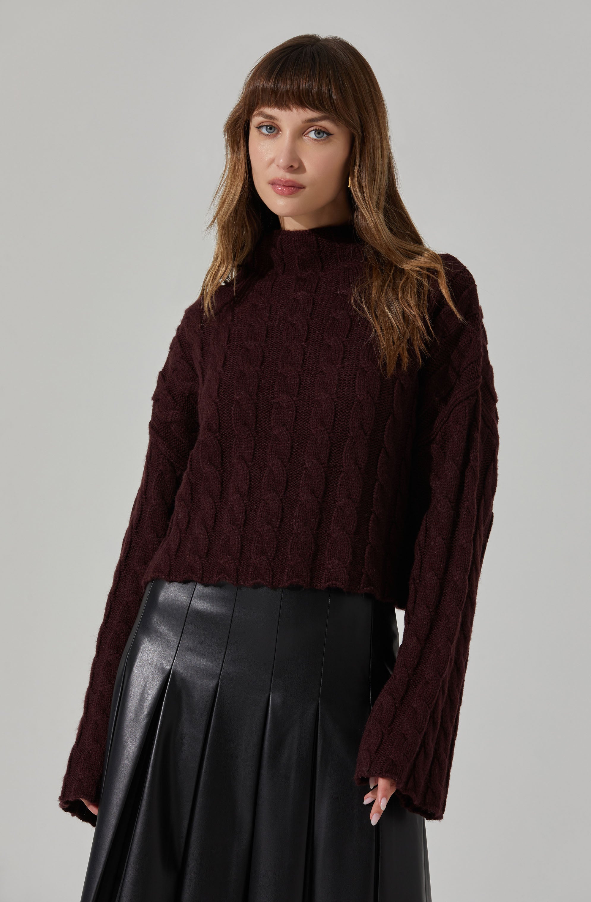A woman with light skin and long, wavy brown hair with bangs is wearing a dark burgundy, cable-knit sweater with a high neckline and long sleeves. She pairs it with a black, high-waisted, pleated leather skirt.