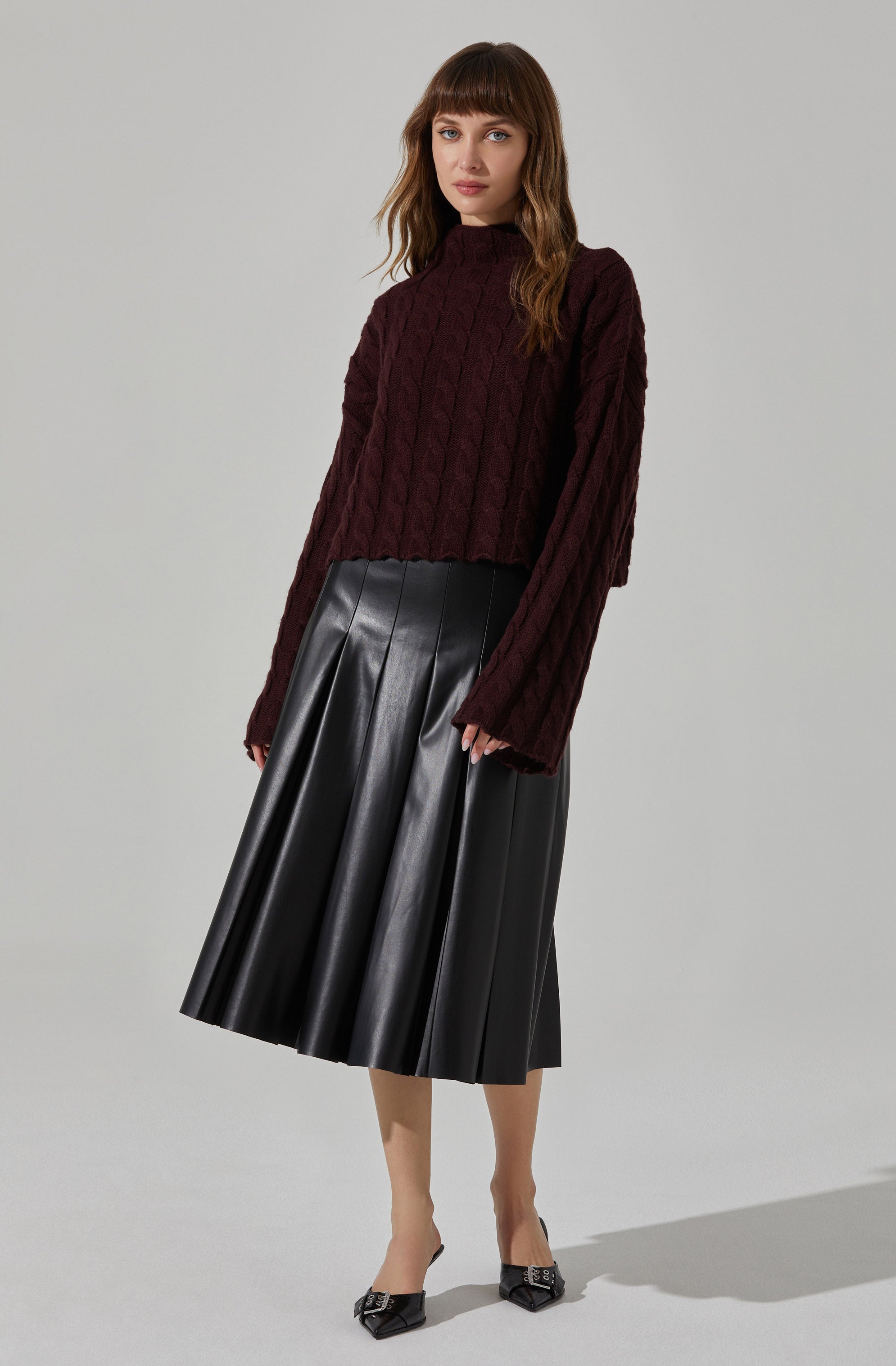 A woman is wearing a dark burgundy, cable-knit sweater with a high neckline and long sleeves. She is also wearing a black, shiny, pleated midi skirt and black pointed-toe heels with ankle straps.