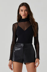 A woman is wearing a black, sheer long-sleeve turtleneck top with a solid black lining underneath. She pairs it with high-waisted black leather shorts.