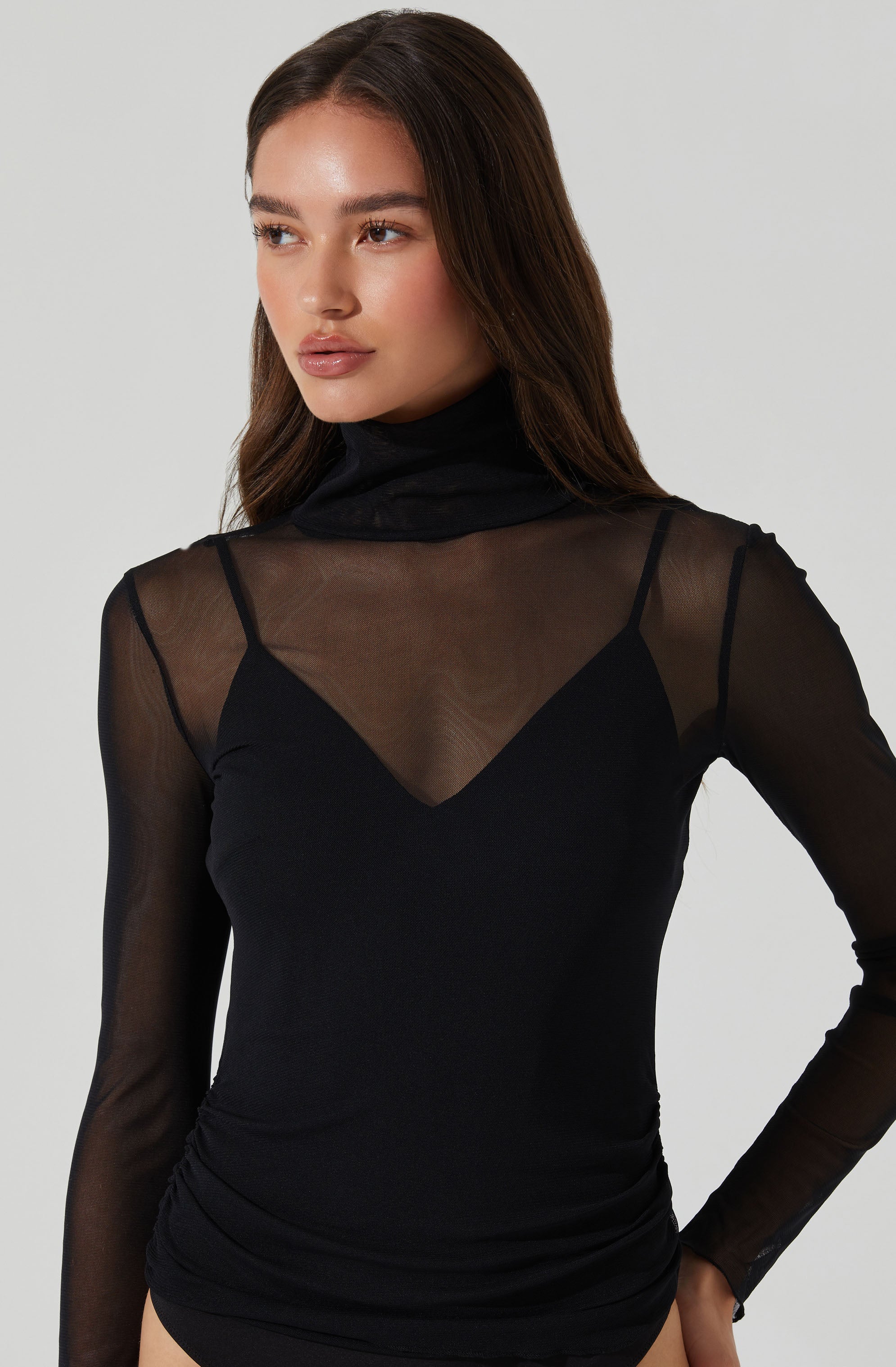 A woman is wearing a black top with a sheer, long-sleeved overlay. The overlay has a high turtleneck collar and is made of a see-through, lightweight fabric. The top underneath has a V-shaped neckline. The shirt fits snugly and is gathered slightly at the waist.