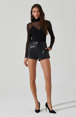 A woman is wearing a black, long-sleeved, sheer turtleneck top with a solid black bra visible underneath. She pairs it with high-waisted black leather shorts and black high-heeled shoes.