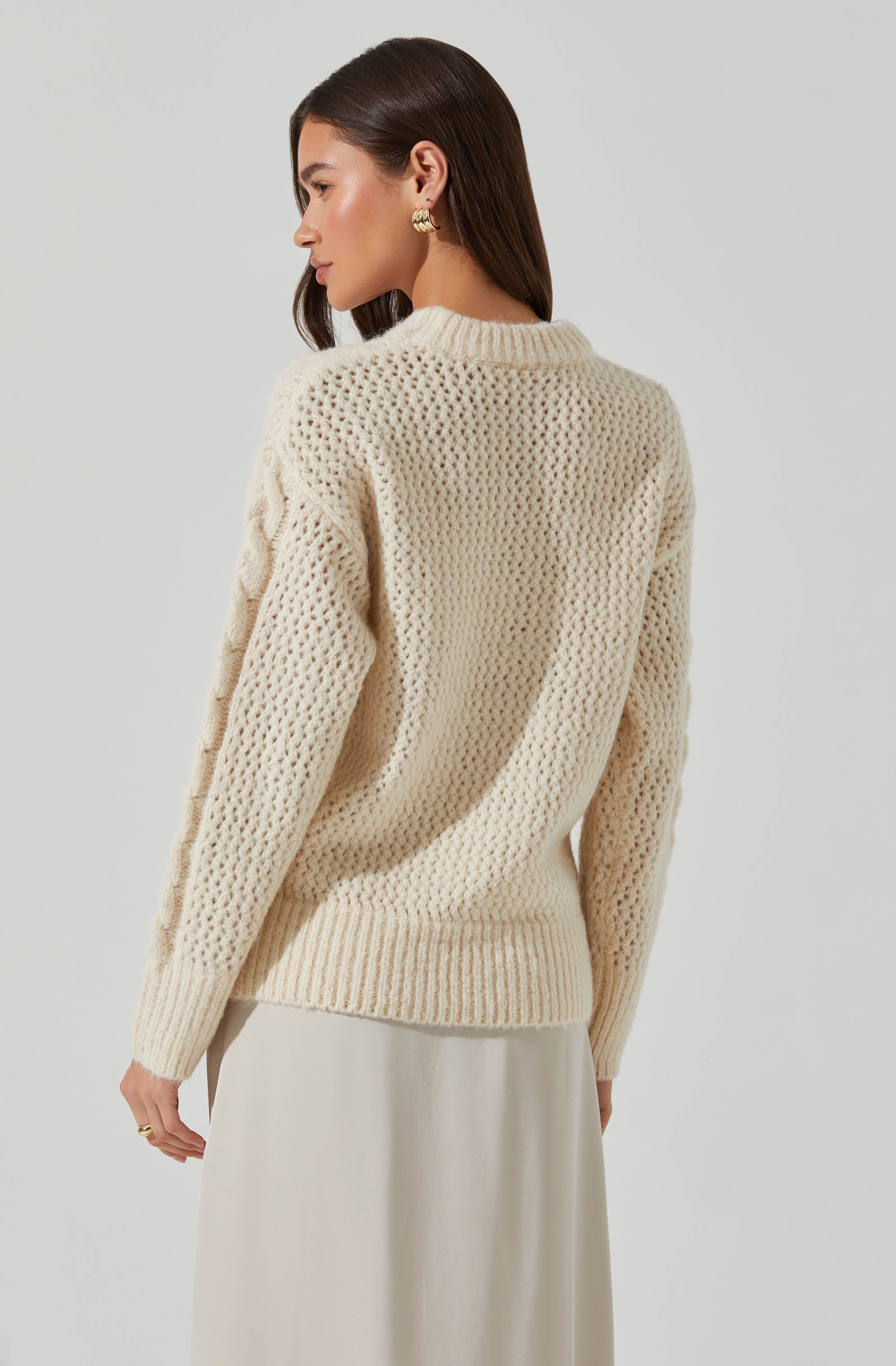 A woman is wearing a cream-colored, chunky knit sweater with a textured, open weave pattern. The sweater has a ribbed neckline, cuffs, and hem. She is also wearing a light-colored skirt.