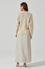 A woman is wearing a chunky, cream-colored knit sweater with a textured pattern and ribbed hem and cuffs. She is also dressed in a long, flowing cream-colored skirt and cream high-heeled shoes.