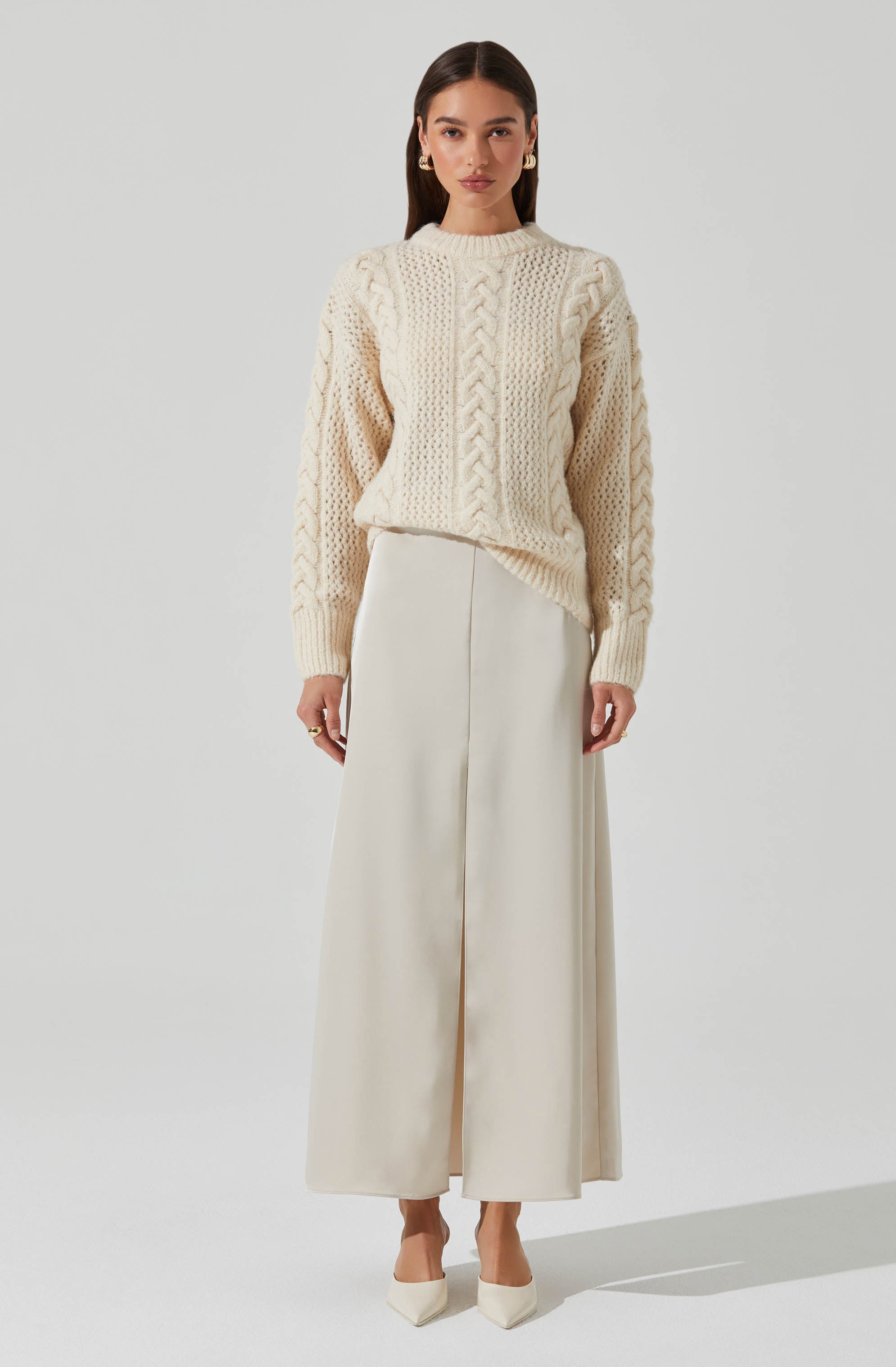 A woman is wearing an oversized cream-colored cable knit sweater with textured patterns and a high neckline. She pairs it with a long, flowing cream-colored skirt that reaches her ankles. She completes her outfit with matching cream heels.