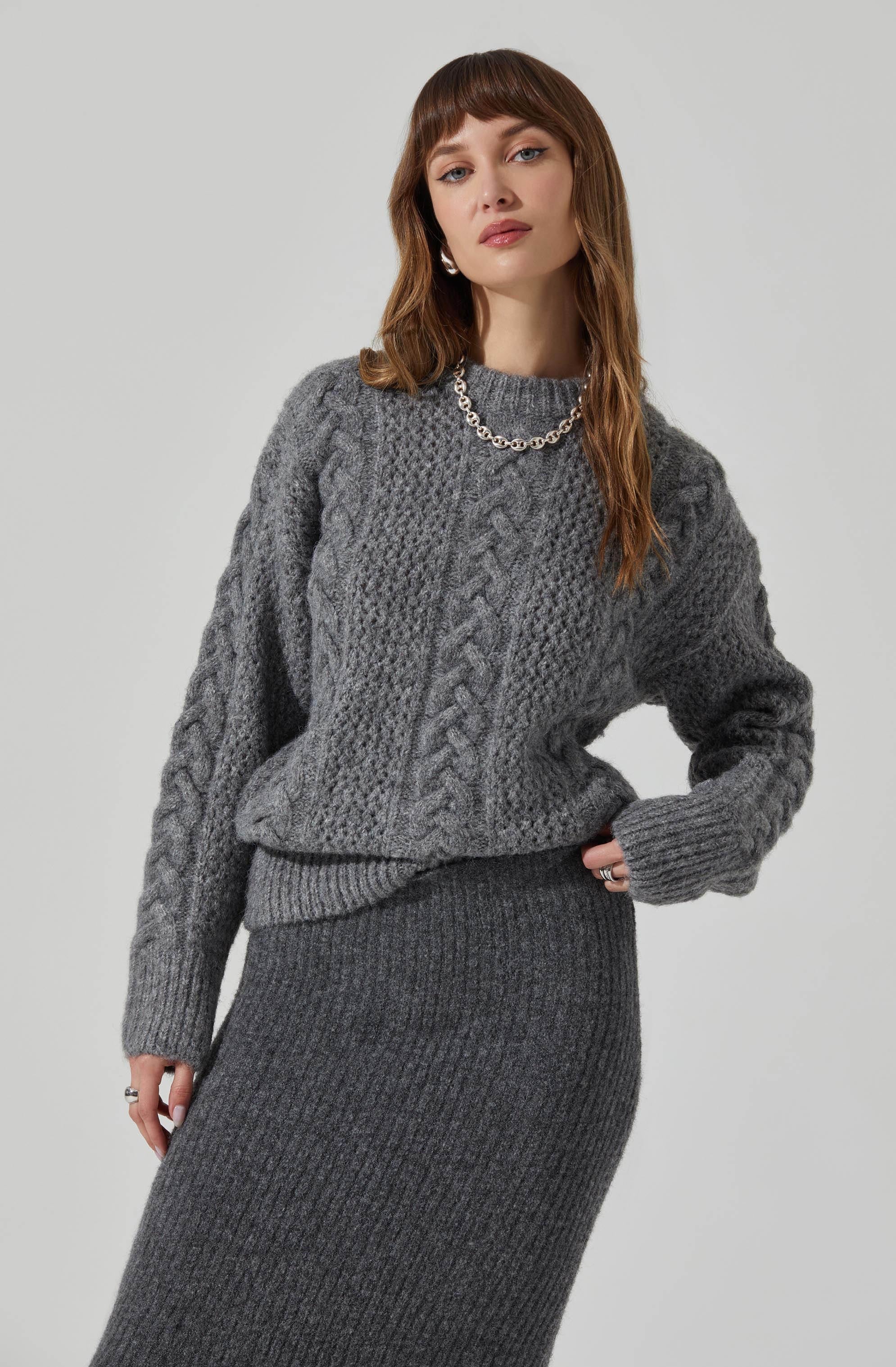 A woman is wearing a gray cable-knit sweater with a crew neckline and long sleeves. She accessorized with a chunky silver chain necklace. She is also wearing a matching high-waisted, ribbed gray skirt.