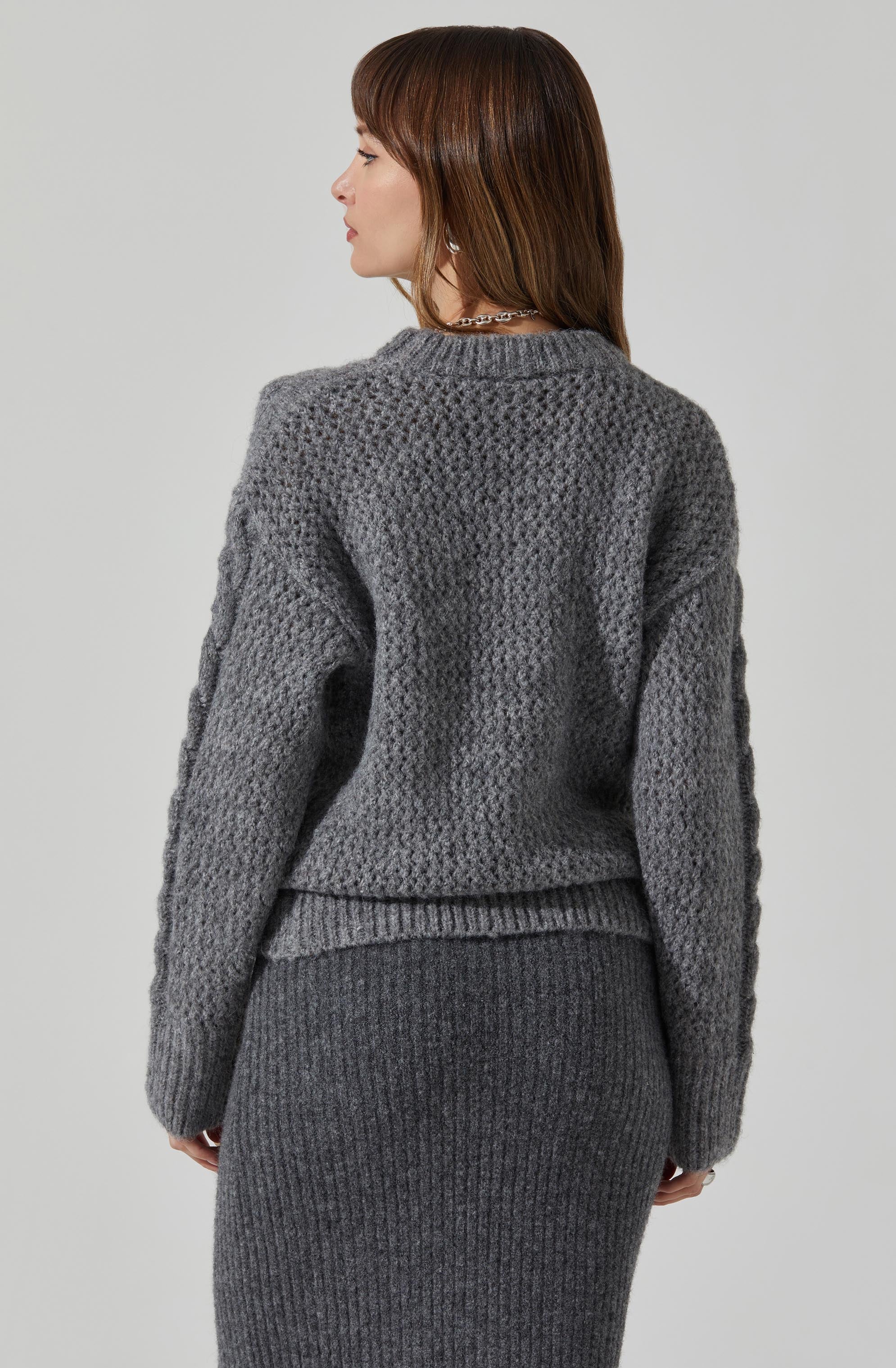 A person is wearing a chunky, textured grey knit sweater with a crew neckline and long sleeves. The sweater has a slightly loose fit and features a waffle pattern throughout. They are also wearing a matching fitted grey ribbed knit skirt that extends below the knees.
