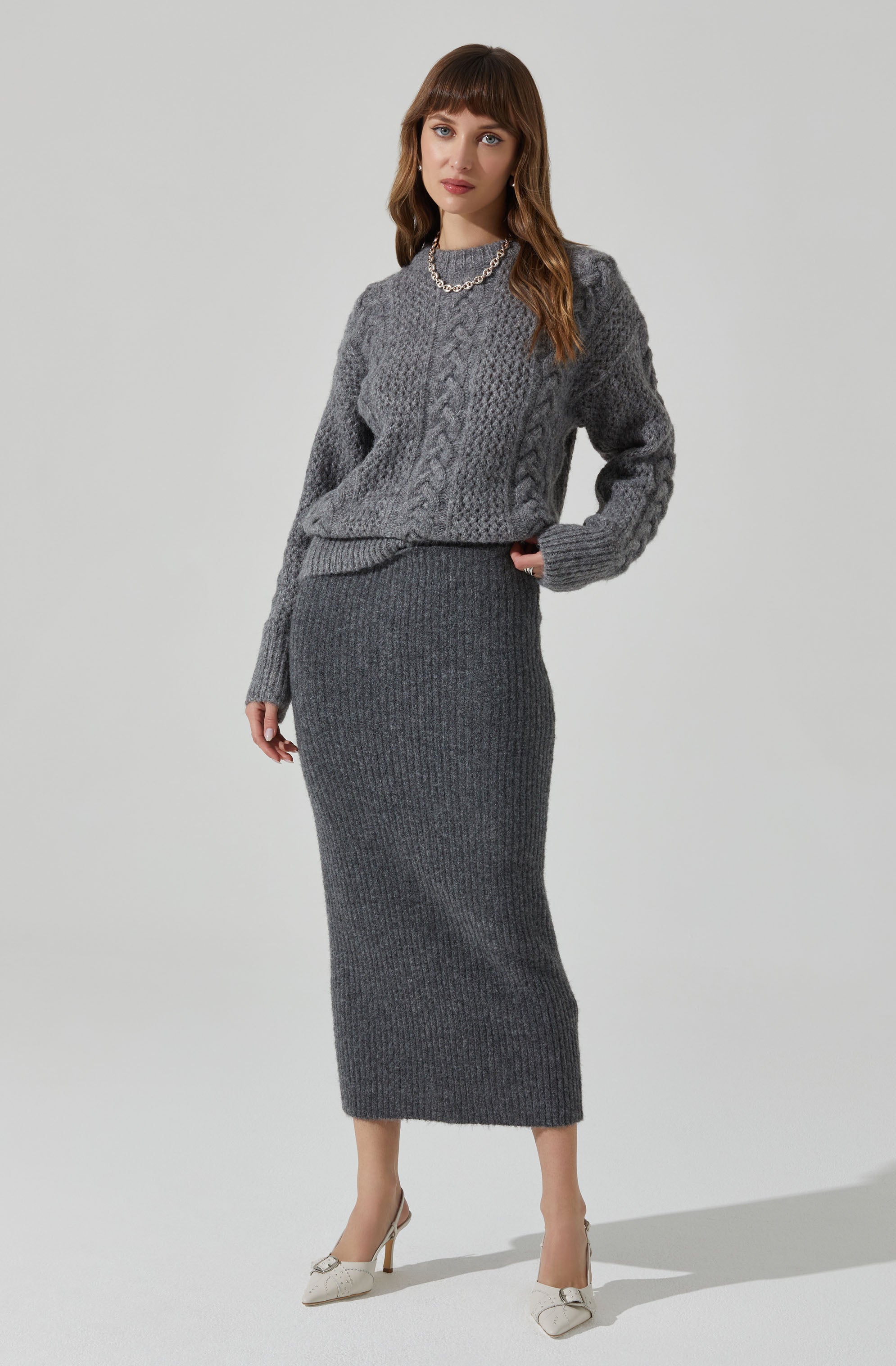 A woman is wearing a gray, chunky knit crewneck sweater with a cable knit pattern and long sleeves. She pairs it with a high-waisted, ribbed gray knit maxi skirt that extends to her ankles. She completes the outfit with cream-colored high heels featuring decorative bows, and she is wearing a pearl necklace.