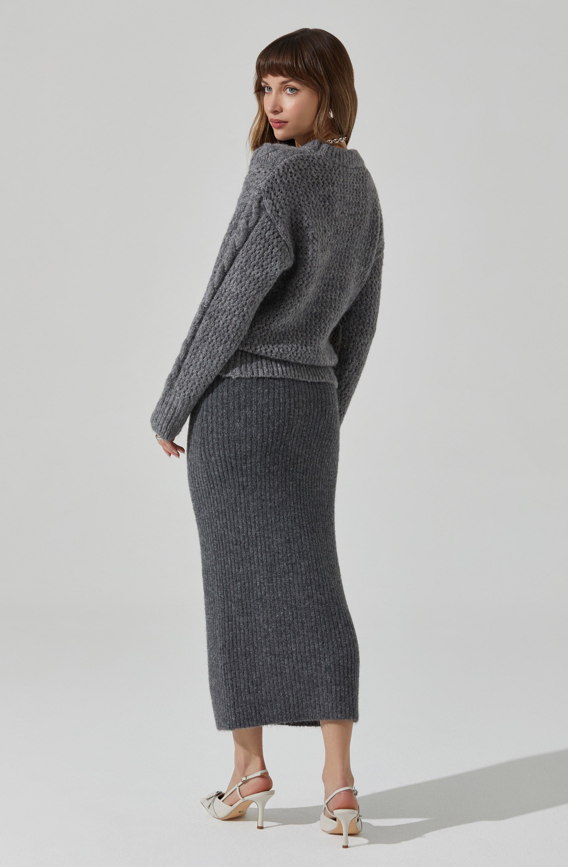 A woman is wearing a cozy, chunky-knit gray sweater with a textured pattern and long sleeves. She is also wearing a matching high-waisted, ribbed knit gray skirt that extends to her ankles. She has on white high-heeled sandals with ankle straps.