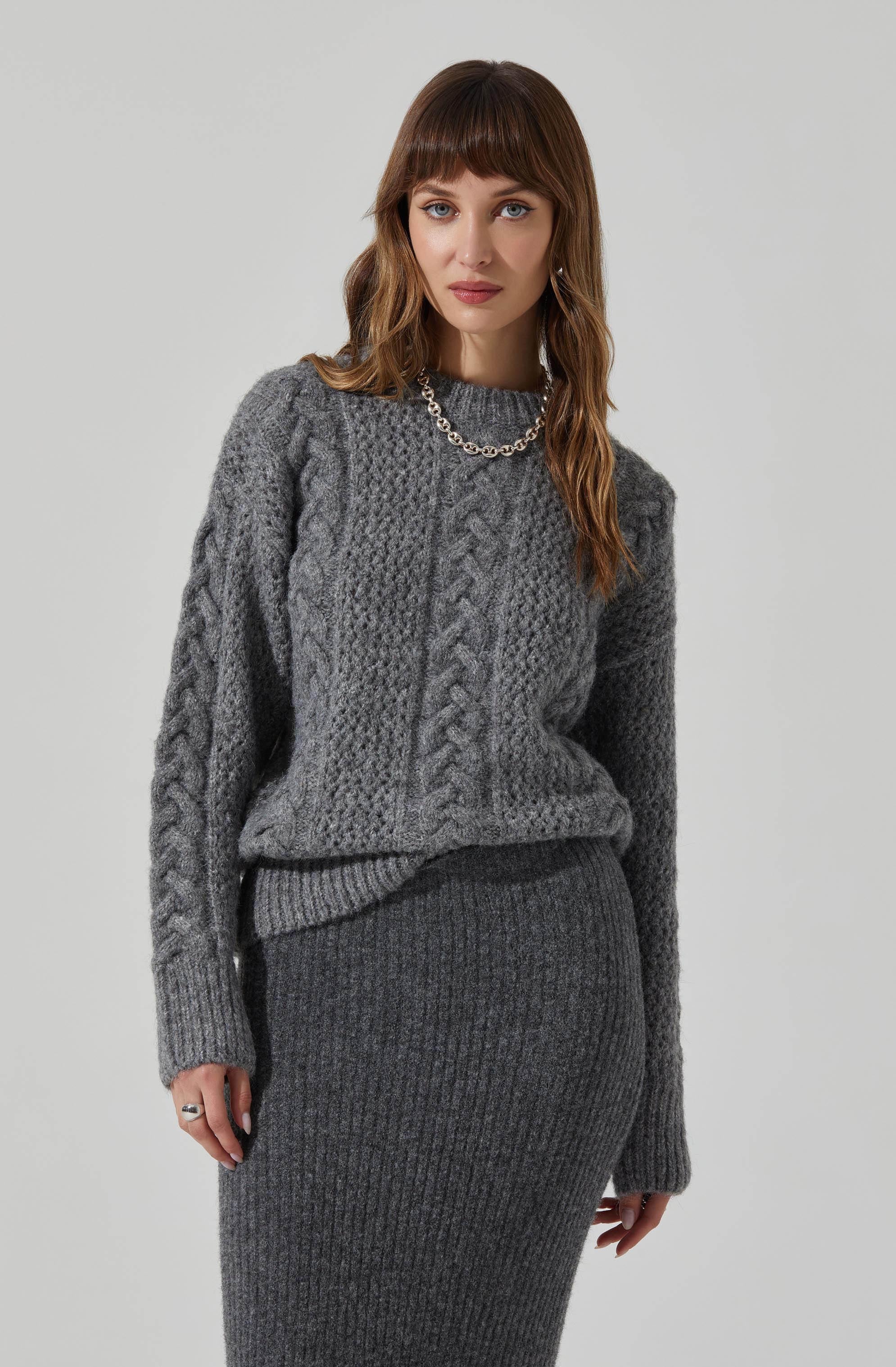 A woman is wearing a chunky knit sweater with cable-knit patterns in various sections, featuring a crew neckline and long sleeves. She pairs it with a high-waisted, ribbed knit skirt, both in shades of gray. She accessorizes with a silver chain necklace and wears rings on her fingers.