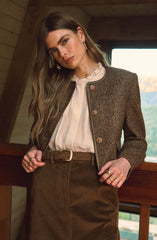 A woman is wearing a textured, dark brown, collarless jacket with gold buttons, and a light-colored blouse with a ruffled neckline underneath. She has on high-waisted brown pants with a belt. She is accessorized with multiple rings, earrings, and has long, wavy hair.