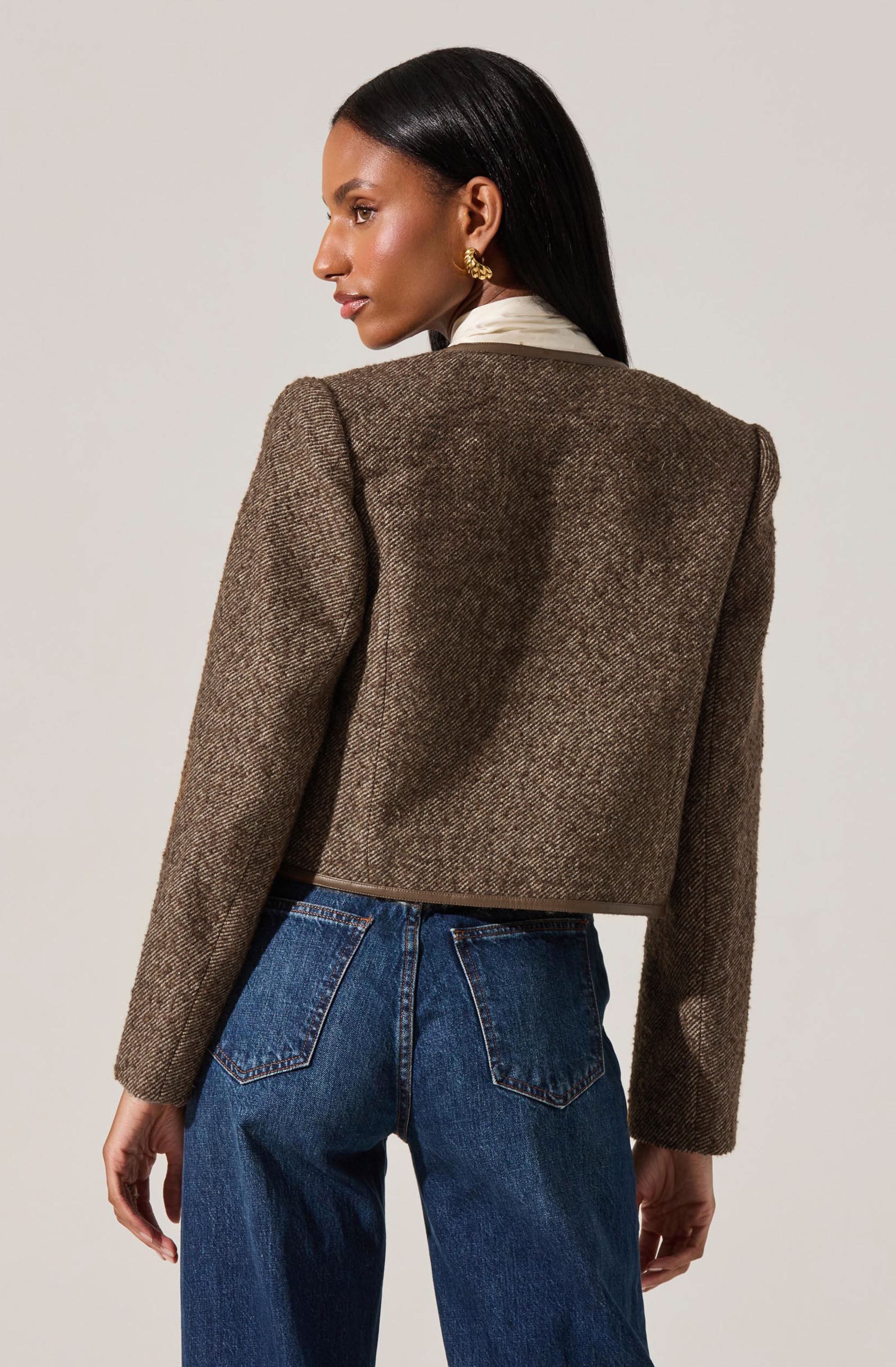 A woman is wearing a brown, textured, cropped blazer with slightly padded shoulders, paired with dark blue high-waisted jeans. The blazer has a subtle herringbone pattern and a structured fit. She also has on a white shirt underneath the blazer and gold earrings.