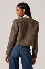 A woman is wearing a brown, textured, cropped blazer with slightly padded shoulders, paired with dark blue high-waisted jeans. The blazer has a subtle herringbone pattern and a structured fit. She also has on a white shirt underneath the blazer and gold earrings.
