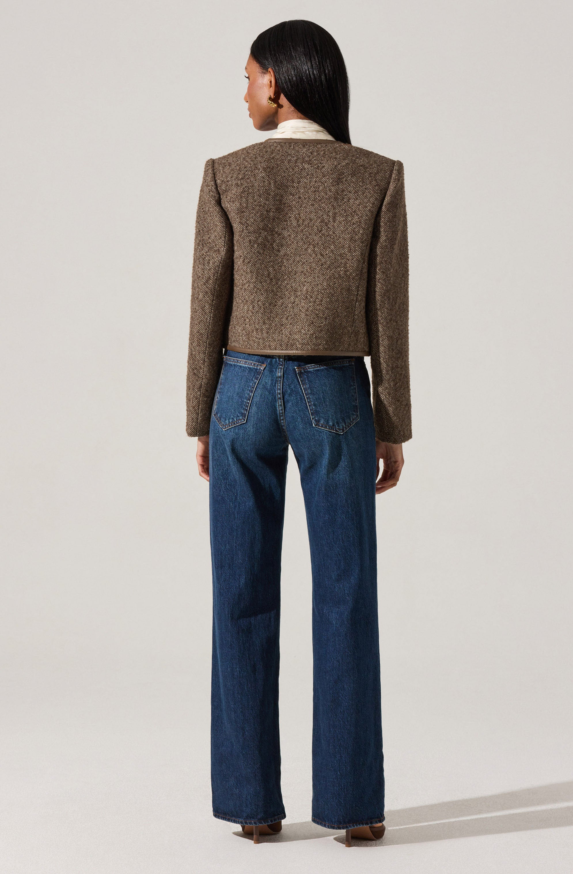 A woman is wearing a beige, textured, cropped blazer with long sleeves over a white collared shirt. She pairs this with high-waisted, straight-leg blue jeans.