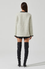 A woman is wearing a cream-colored, textured boucle jacket with black trim around the collar, cuffs, and hem. She is also wearing black shorts and over-the-knee black leather boots with high heels.