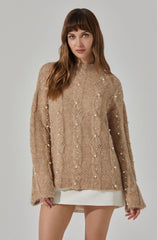A woman is wearing a loose-fitting, beige cable-knit sweater embellished with vertical rows of small, white pearls. The sweater has a high neckline, long sleeves, and a relaxed fit that extends past her hips. She pairs it with a short, white skirt that is barely visible below the hem of the sweater.