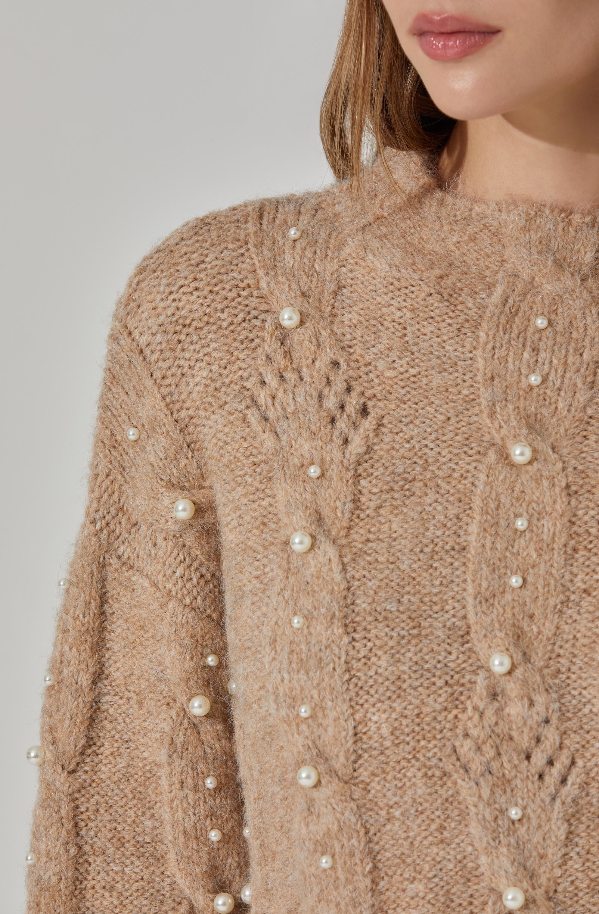 A close-up of a woman wearing a beige knitted sweater embellished with small, round pearl accents scattered across the fabric. The sweater has a textured pattern and a high neckline.