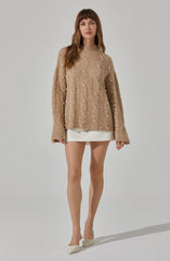 A woman is wearing a loose, oversized beige cable-knit sweater decorated with small white pearls scattered throughout. She pairs it with a short, cream-colored skirt and beige high-heeled shoes.