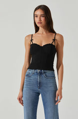 A woman is wearing a black sleeveless top with thin spaghetti straps that have small bows tied at the shoulders. The top features a structured, possibly corset-style design with seam details and a slight ruffle trim along the neckline. She pairs the top with high-waisted blue jeans that have a classic fit and a button and zip closure at the front.
